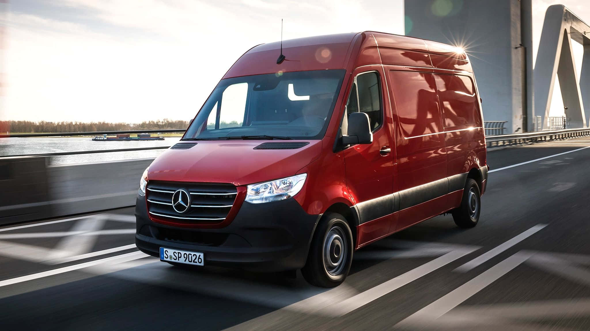 Van 桌布 壁紙 — 一輛時尚的紅色 Mercedes-Benz Sprinter 客貨車在明亮的陽光下行駛在現代化的橋樑上，背景有水和樹木。