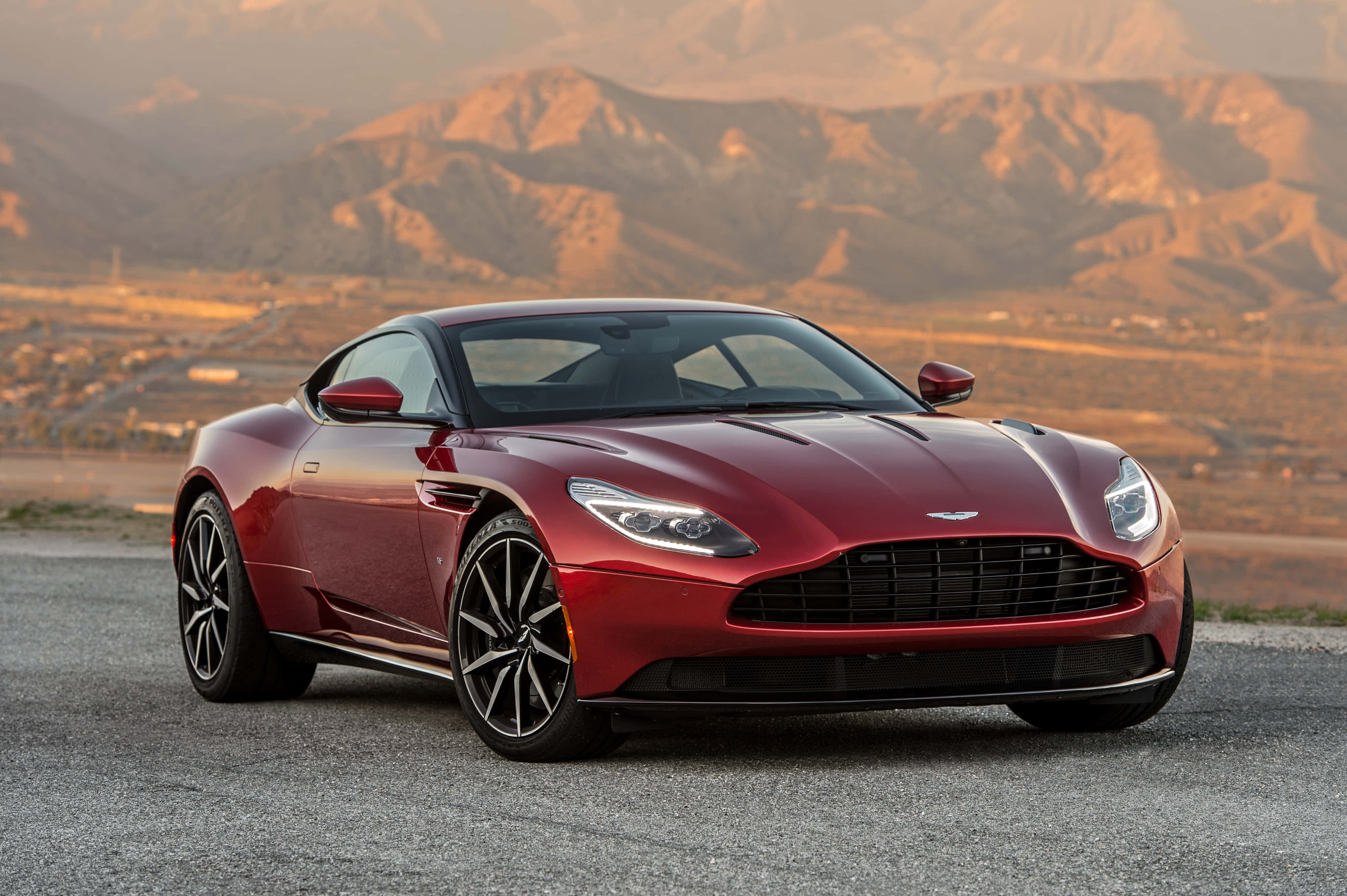 Aston Martin Db11 桌布 壁紙 — 一輛時尚的紅色Aston Martin跑車在碎石路上閃閃發光，背景是陽光明媚的山脈。