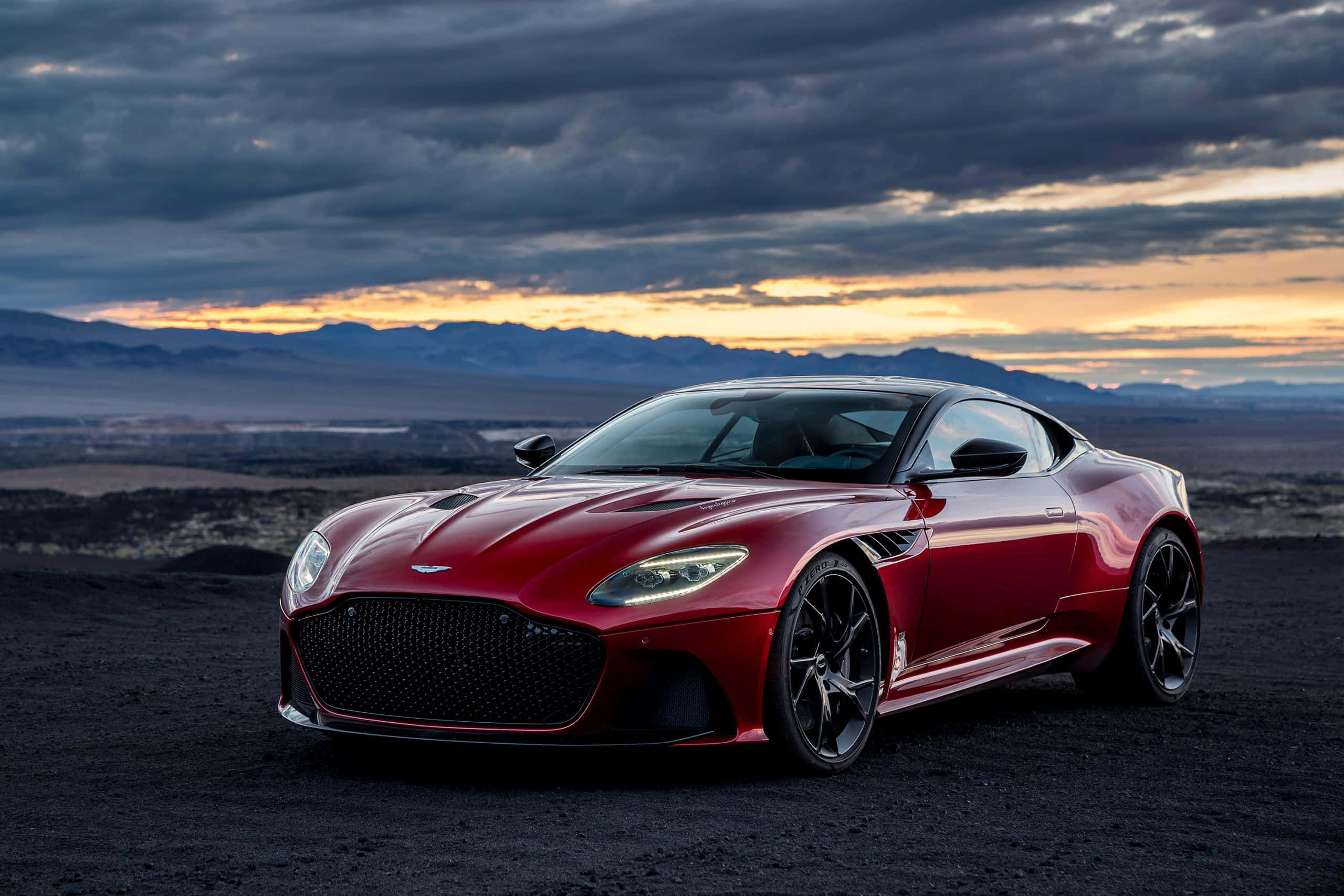 奧斯頓馬丁 桌布 壁紙 — 一輛時尚的紅色Aston Martin跑車在戲劇性的日落天空下閃耀，天空中有烏雲和遠山。