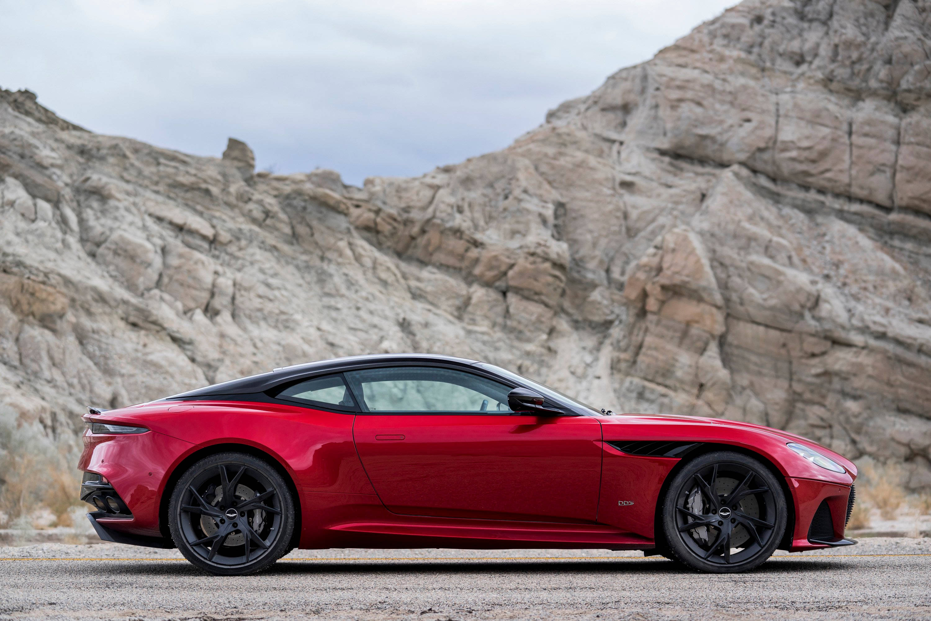 奧斯頓馬丁 桌布 壁紙 — 一輛時尚的紅色Aston Martin跑車，帶有黑色點綴，停泊在碎石路上，背景是崎嶇的岩石懸崖，天空多雲。