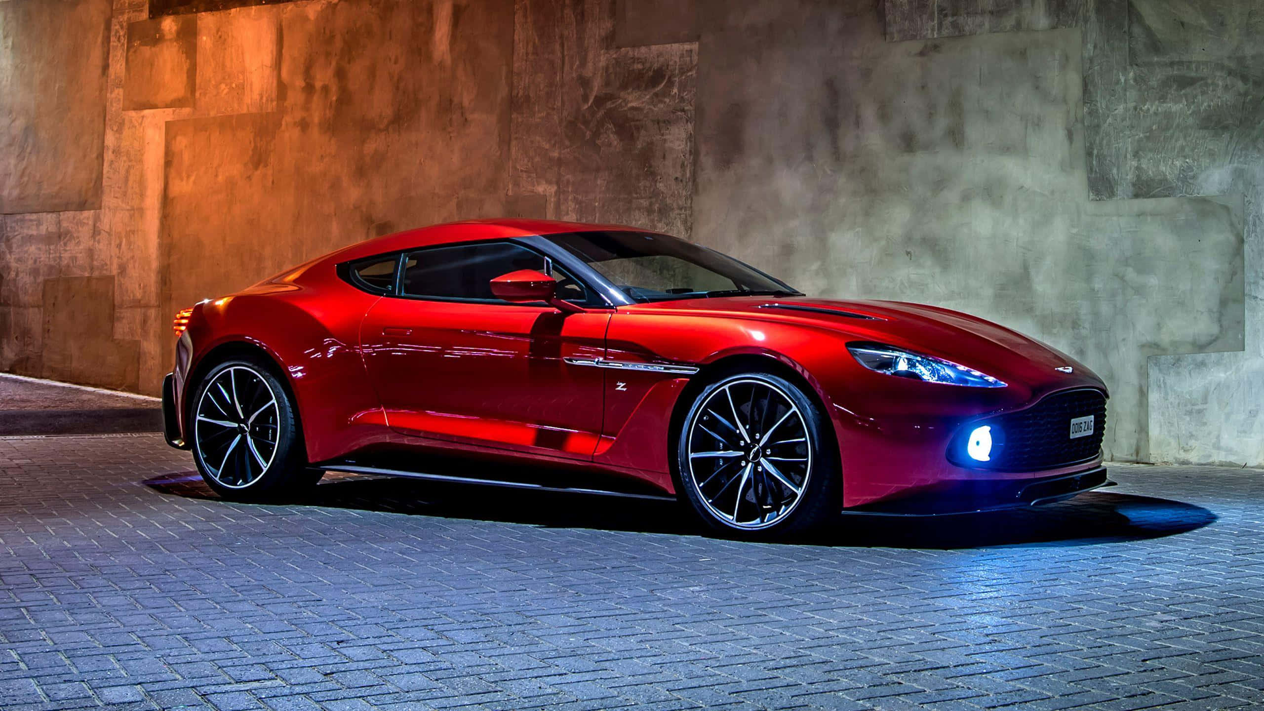 奧斯頓馬丁 桌布 壁紙 — 一輛時尚的紅色Aston Martin跑車在戲劇性燈光下，在鵝卵石街道上以紋理混凝土牆為背景閃閃發光。