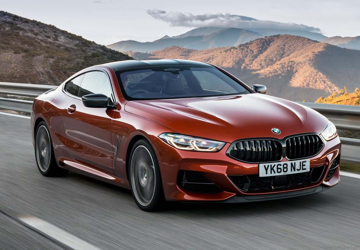 Bmw 8 Series 桌布 壁紙 — 一輛時尚的紅色BMW雙門跑車在崎嶇山丘和柔和雲朵的背景中沿著山區高速公路飛馳。