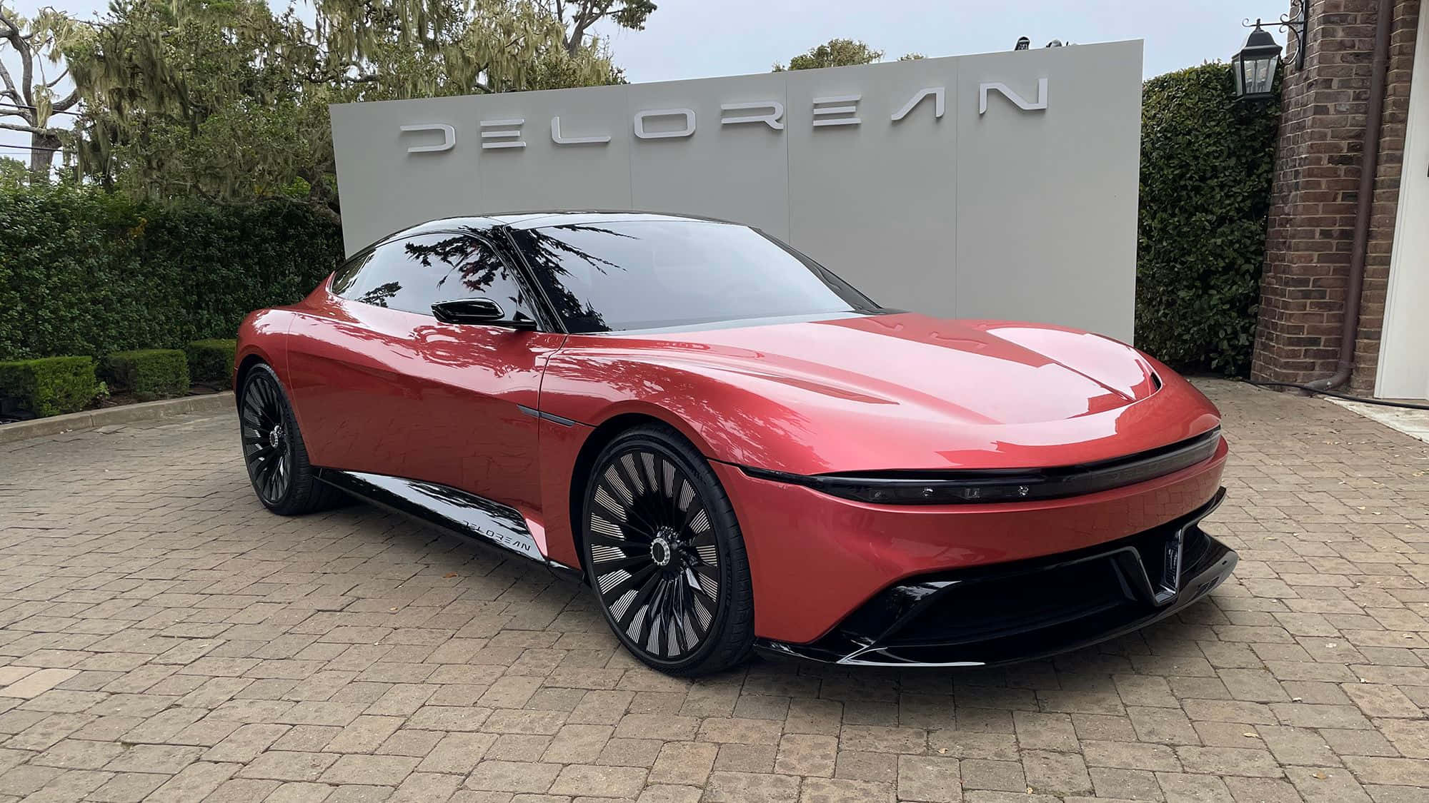 Red Car 桌布 壁紙 — 一輛時尚的紅色DeLorean電動車在日光下閃耀，停在石頭車道上，旁邊是帶有品牌的牆壁。