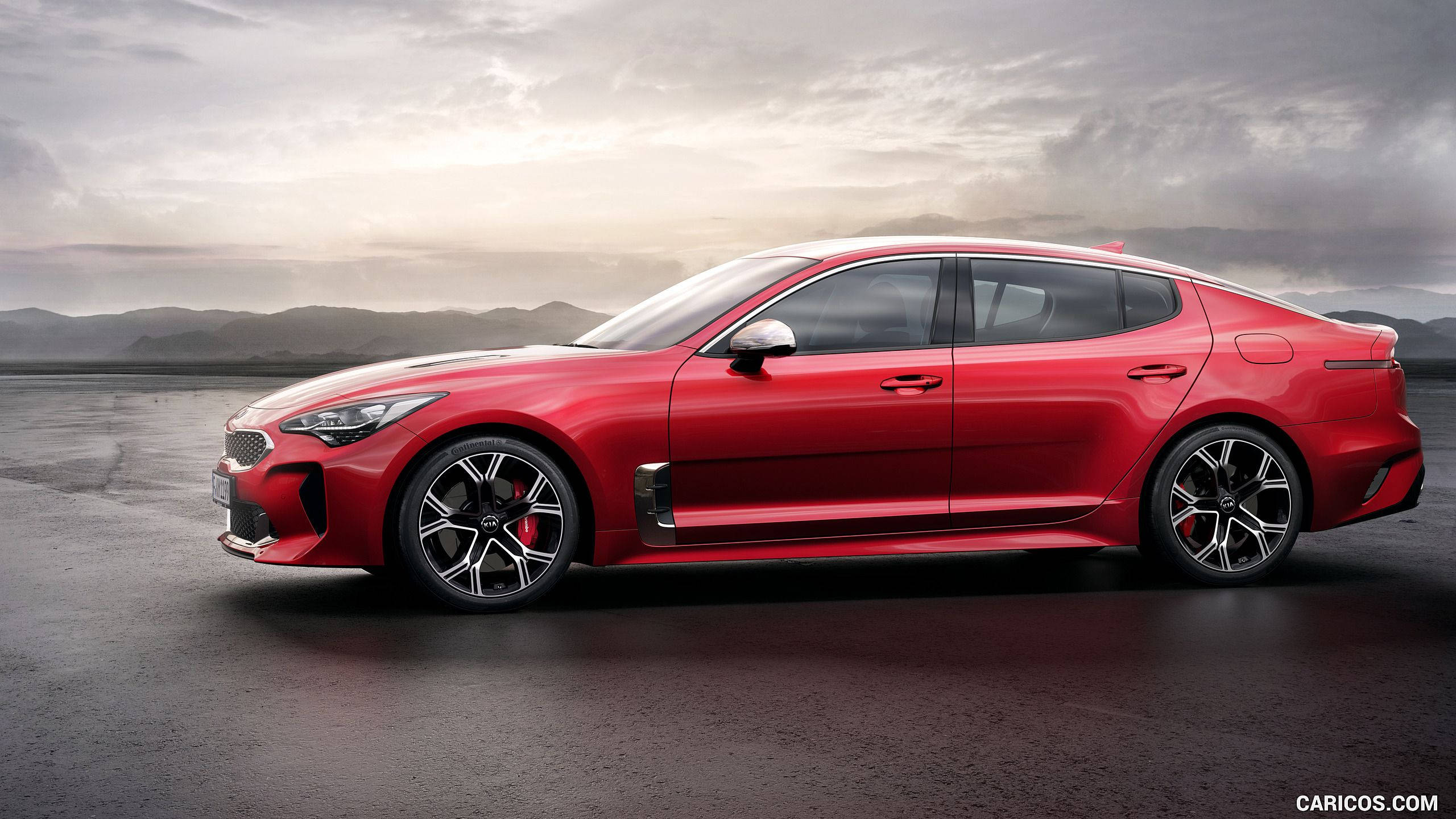 Kia 桌布 壁紙 — 一輛時尚的紅色Kia Stinger運動轎車在陰沉多雲的天空下閃閃發光，背景是遠處的山脈。