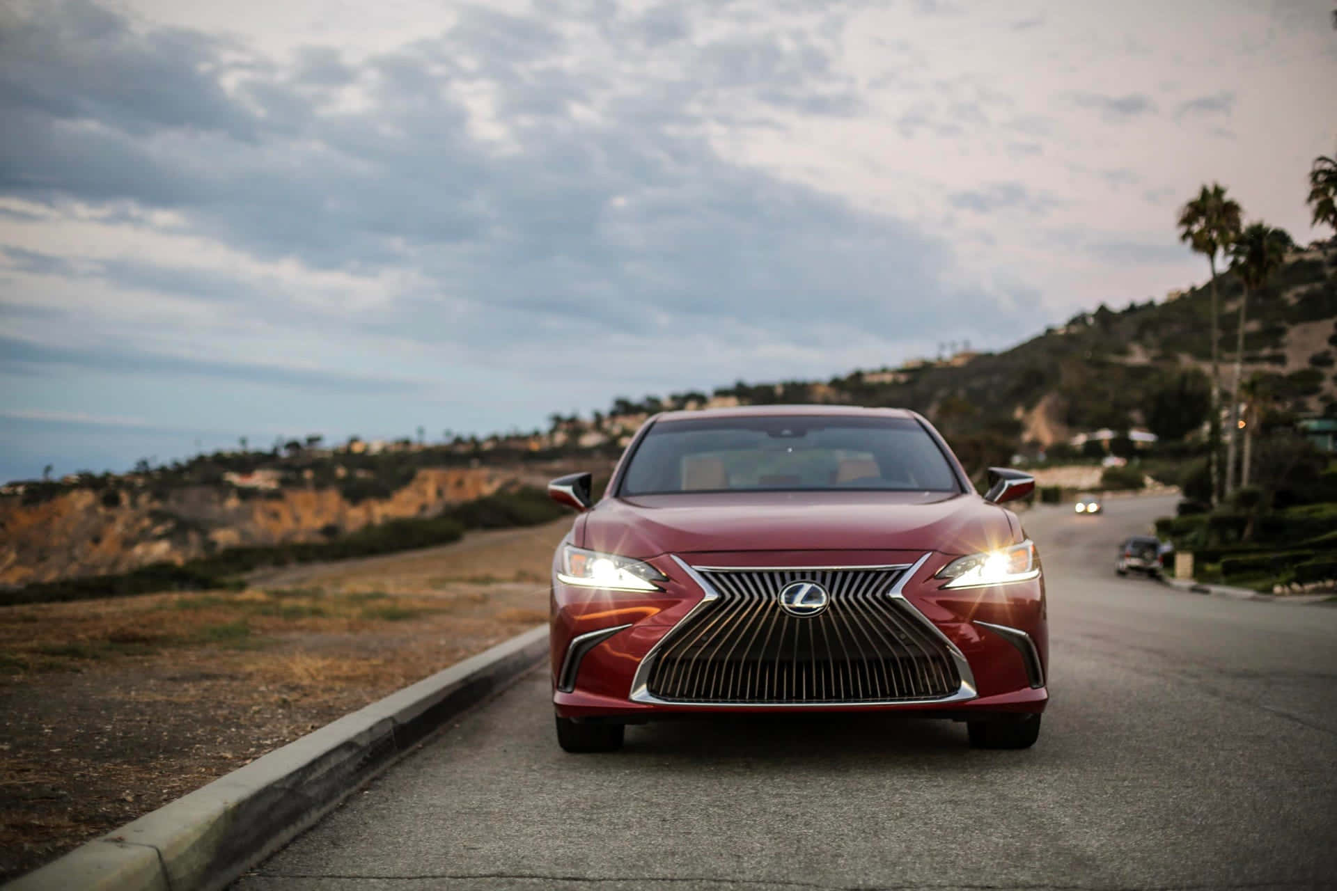 Lexus Es 300h 桌布 壁紙 — 一輛時尚的紅色Lexus轎車在黃昏時分面向海岸道路，背景有棕櫚樹和山丘。