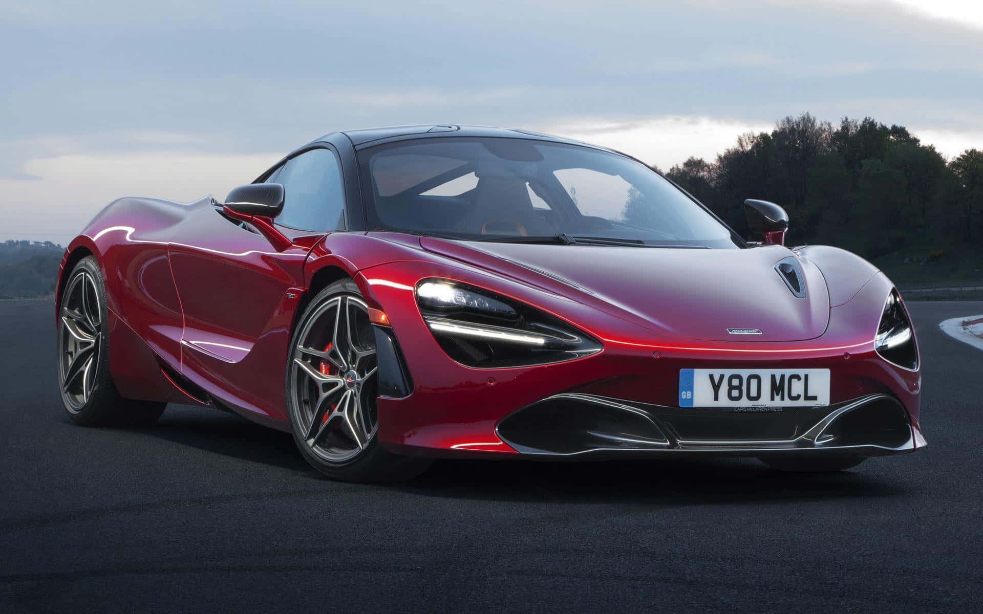 麥拉倫 720S 桌布 壁紙 — 一輛時尚的紅色McLaren 720S跑車在深色瀝青賽道上閃耀，背景有樹木和多雲的天空。