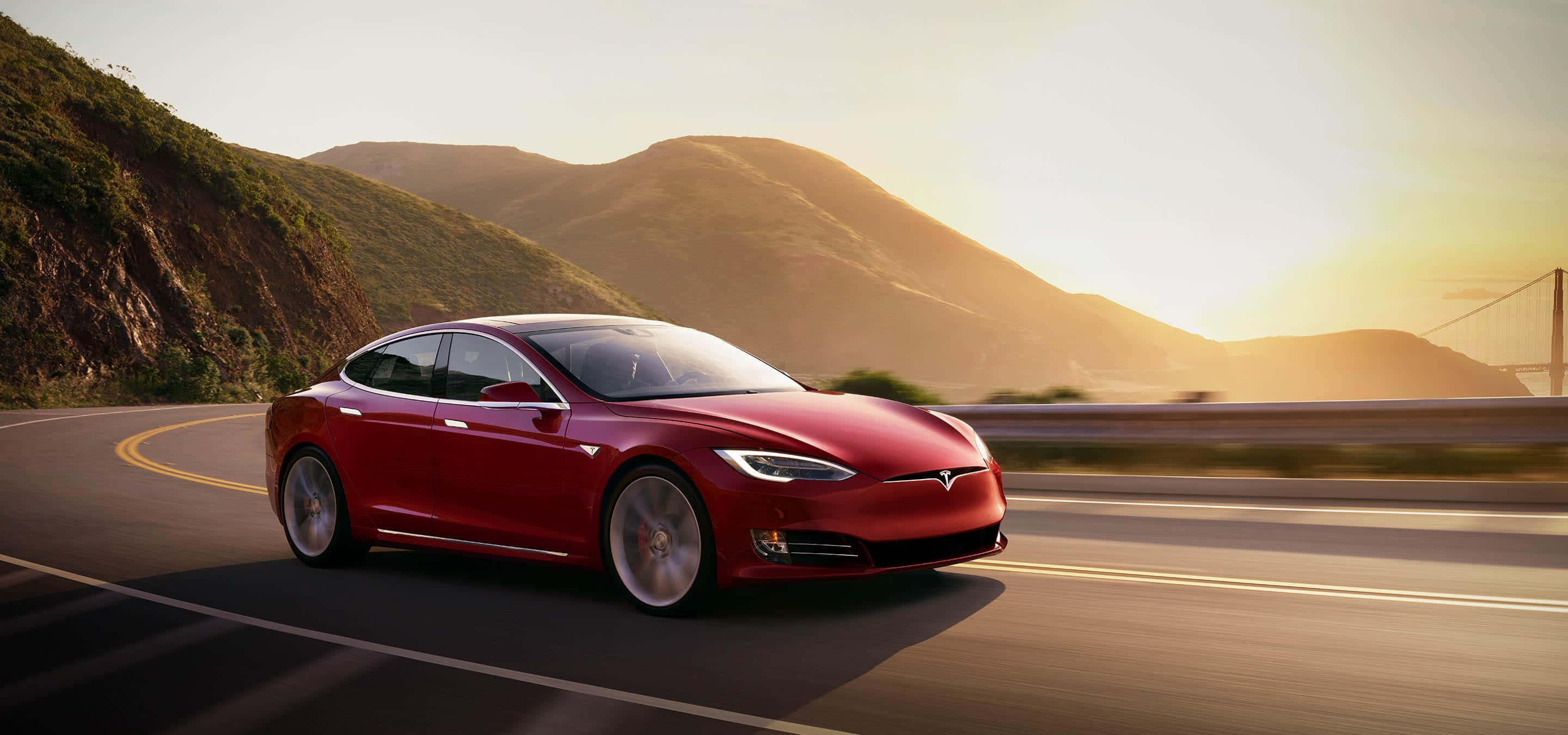 Tesla Model 3 桌布 壁紙 — 一輛時尚的紅色Tesla Model S在金色日落時分沿著蜿蜒的海岸公路行駛，背景有山脈和橋樑。