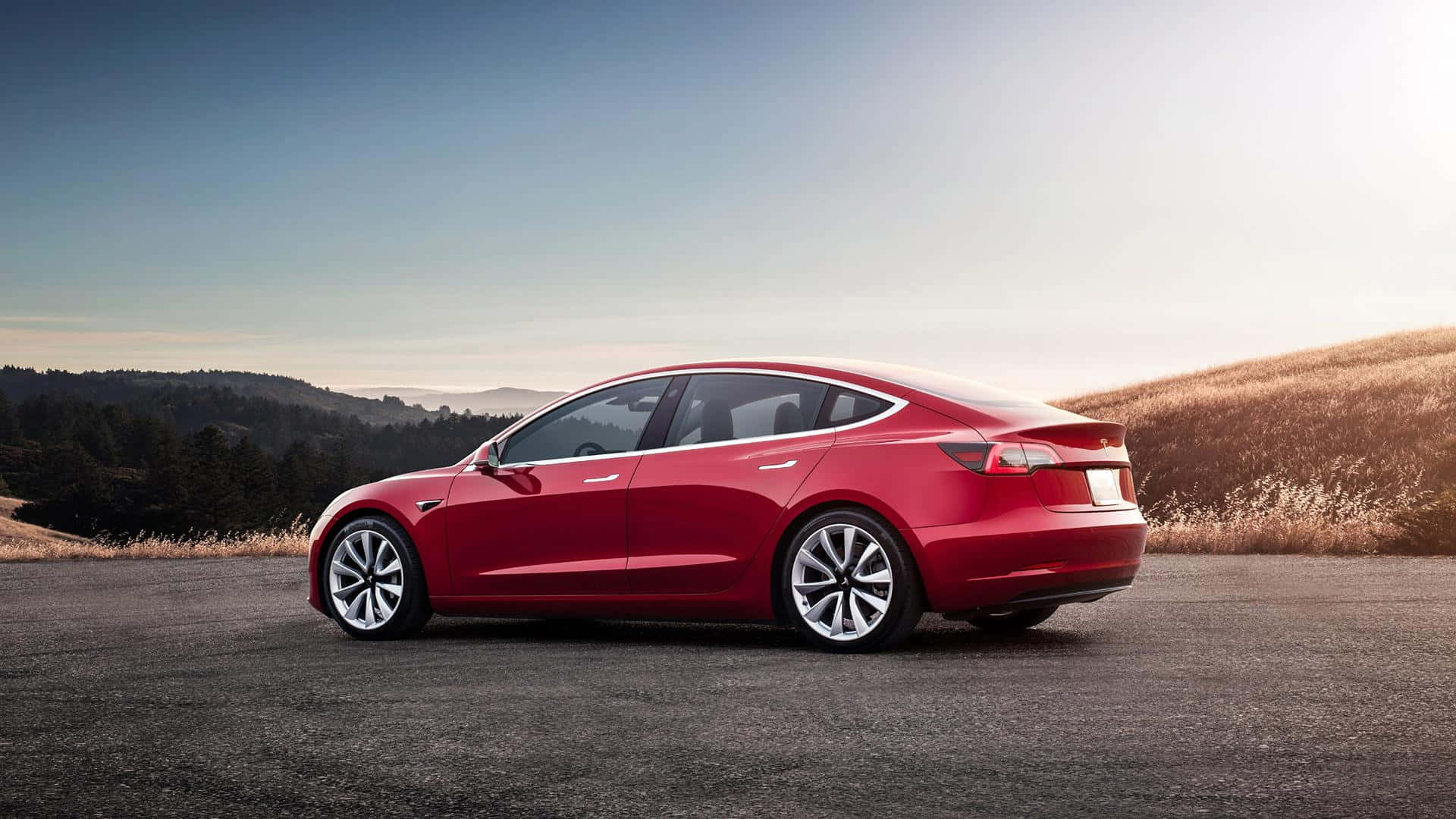 Tesla Model 3 桌布 壁紙 — 一輛時尚的紅色Tesla Model 3停在瀝青路上，背景是起伏的山丘和樹木，在柔和的藍天下，正值黃金時刻。