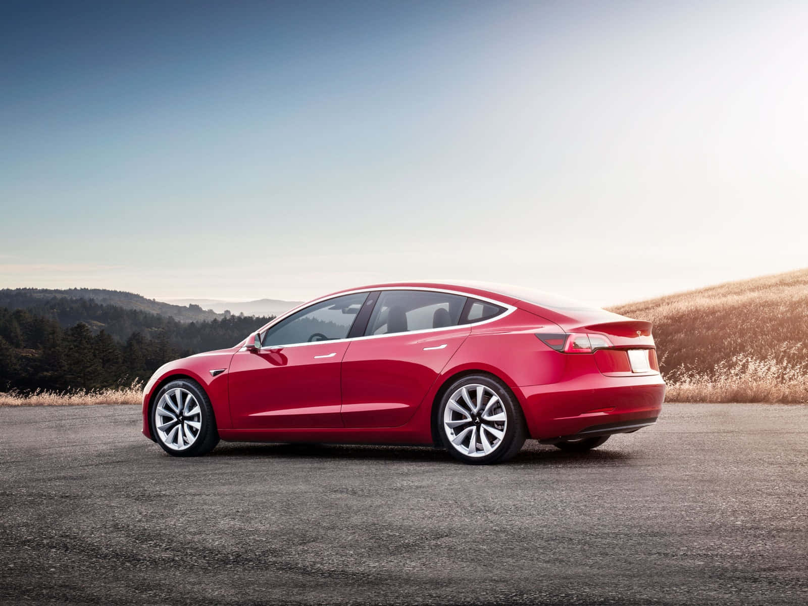 特斯拉 桌布 壁紙 — 一輛時尚的紅色Tesla Model 3停泊在柏油路上，背景是起伏的山丘和樹木，天空晴朗。