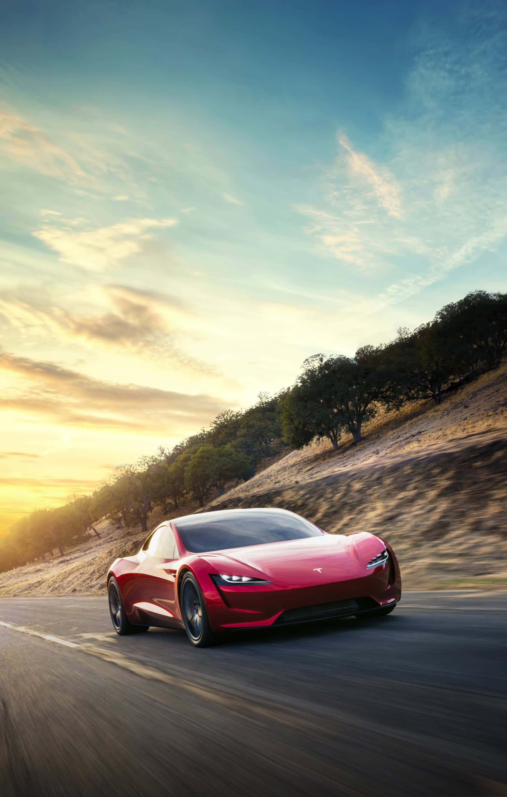 Tesla iPhone 桌布 壁紙 — 一輛時尚的紅色Tesla Roadster在日落時分沿著蜿蜒的道路疾馳，上方有金色的光線和散落的雲朵。