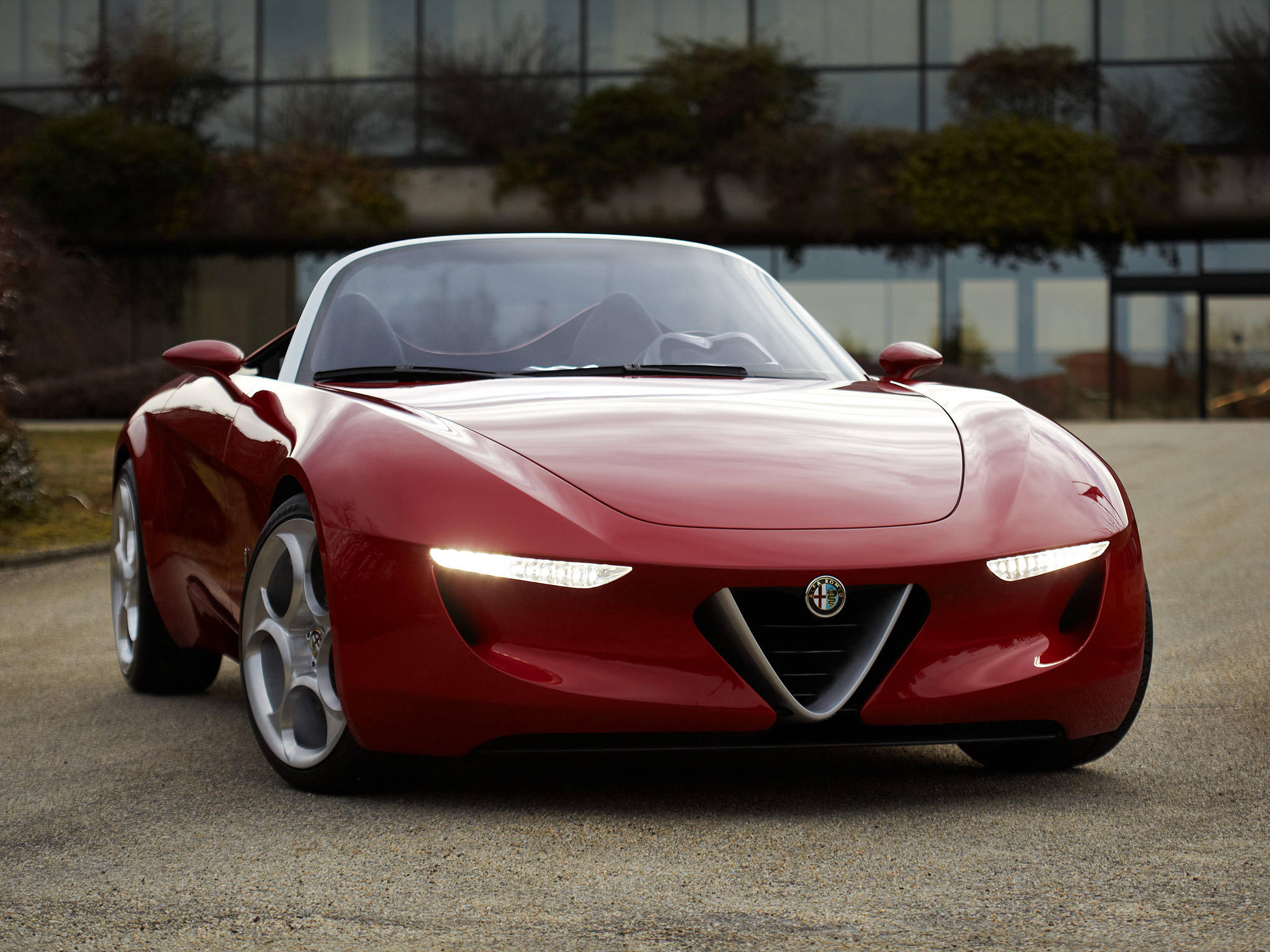 alfa romeo iPhone 桌布 壁紙 — 一輛時尚的紅色愛快羅密歐概念車，帶有發光車頭燈，停在現代玻璃建築前的礫石上。