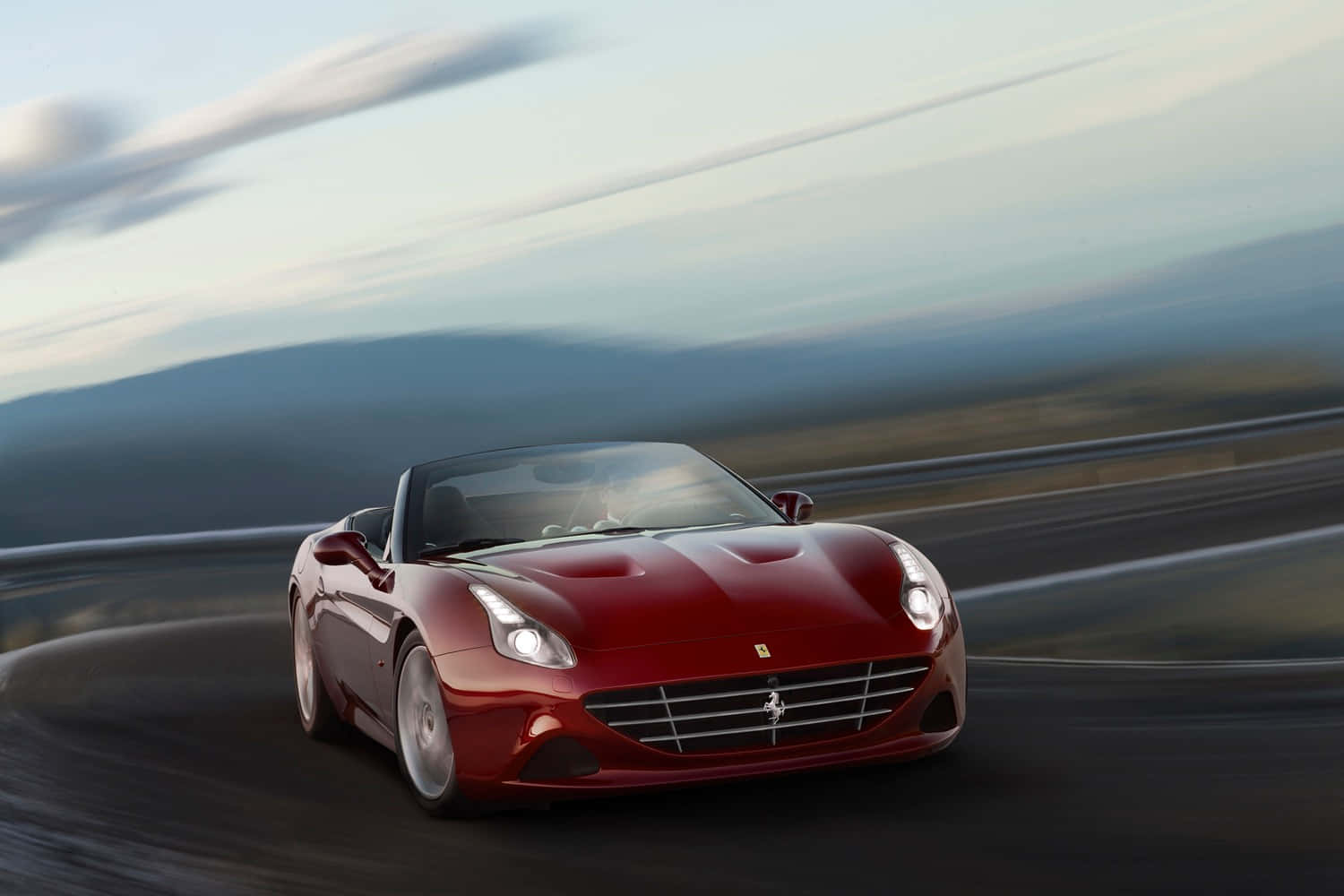 Ferrari California T 桌布 壁紙 — 一輛時尚的紅色法拉利敞篷車在黃昏時分沿著蜿蜒的山路疾馳，帶有模糊的動感和柔和的天空色調。