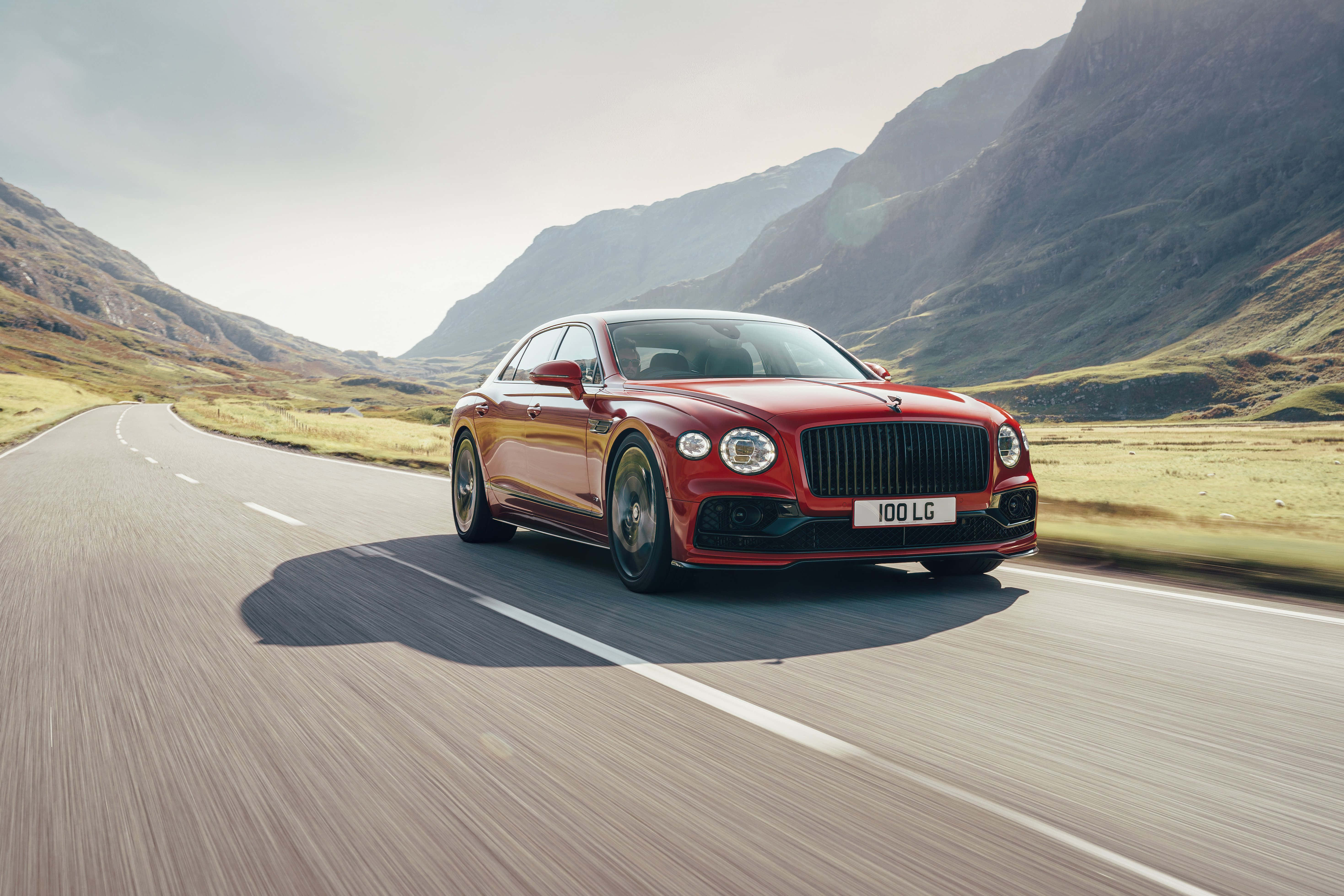 Bentley Flying Spur 桌布 壁紙 — 一輛時尚的紅色賓利飛馳在明亮的天空下沿著蜿蜒的山路飛馳。
