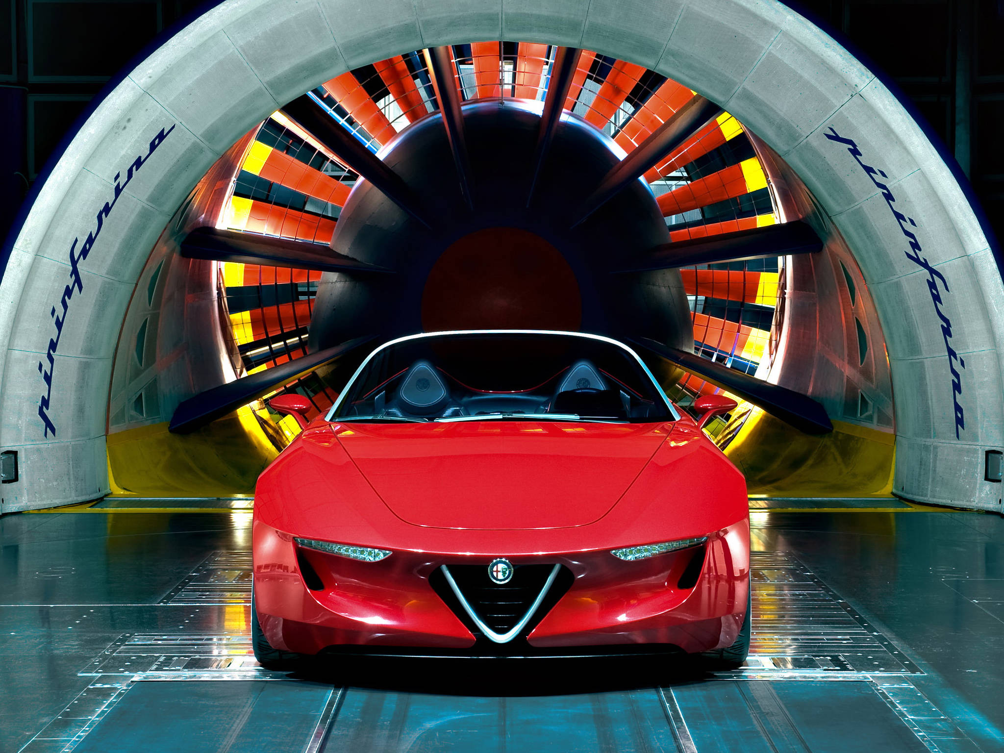 alfa romeo iPhone 桌布 壁紙 — 一輛時尚的紅色跑車位於高科技風洞中央，背後有鮮豔的橙色和黃色葉片旋轉。