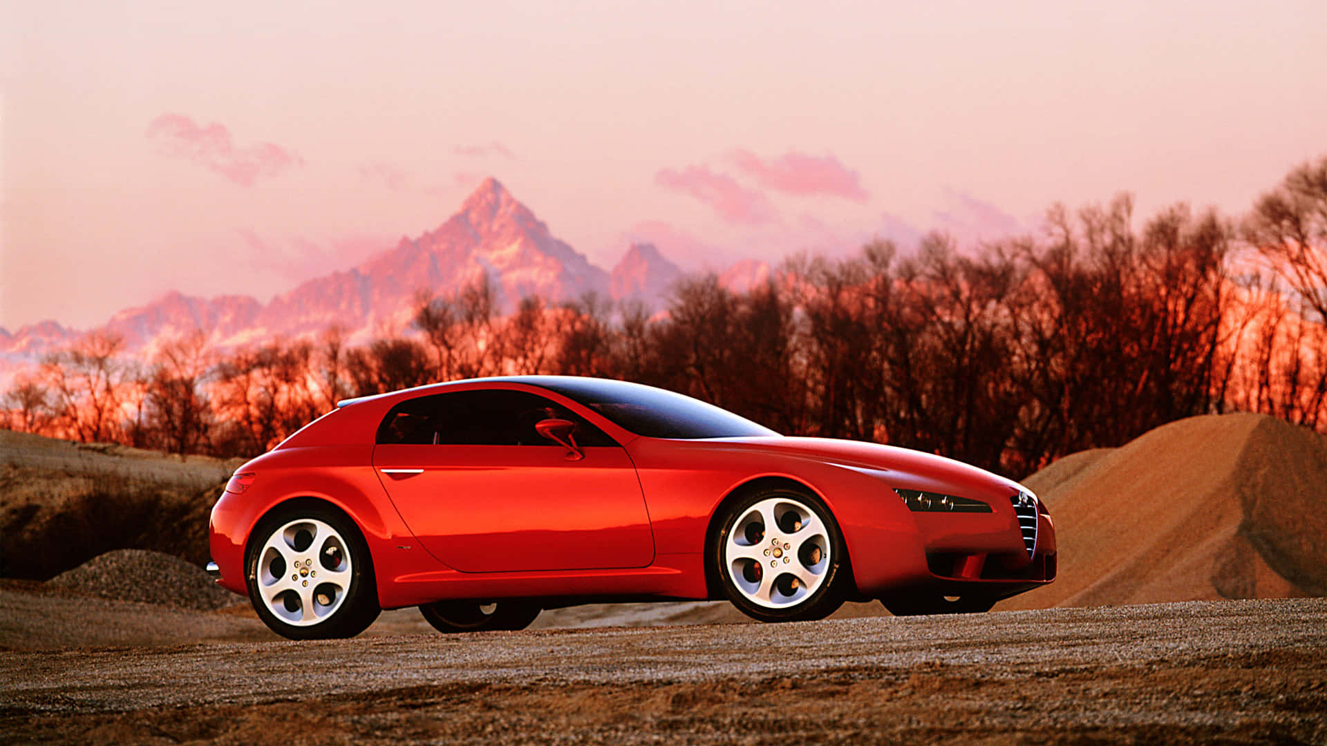 Alfa Romeo Brera 桌布 壁紙 — 一輛時尚的紅色跑車停在碎石上，背景是白雪覆蓋的山脈和光禿禿的樹木在日落色調中閃耀。