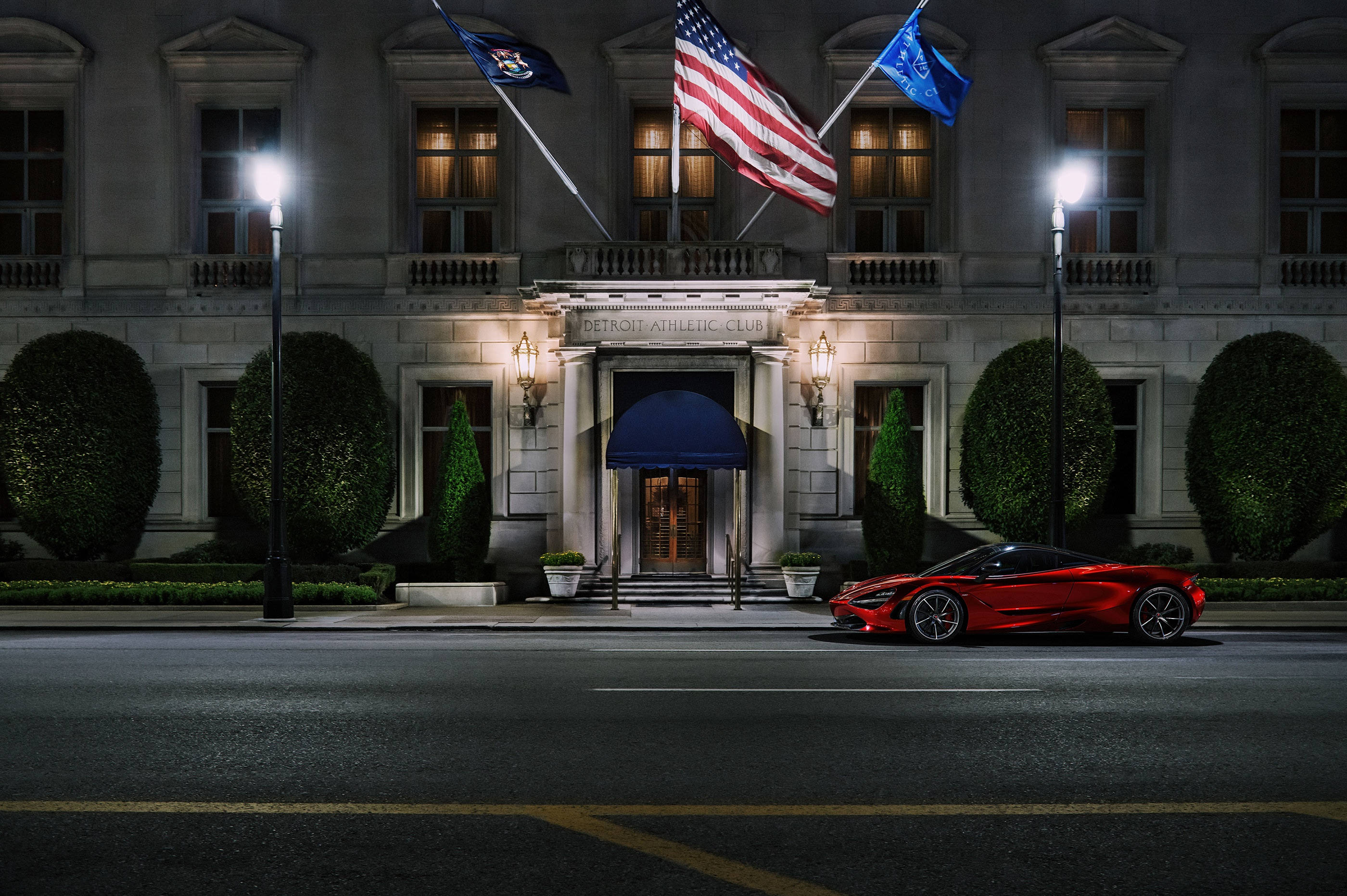 Mclaren 720s 桌布 壁紙 — 一輛時尚的紅色跑車在夜間停在一座宏偉的燈光建築前，建築物有旗幟和修剪整齊的樹籬。