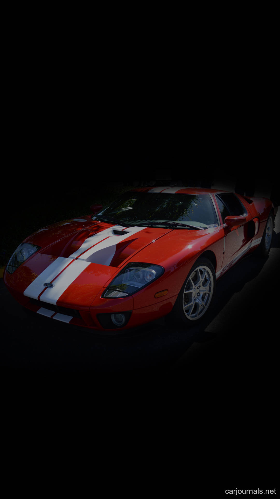 ford iPhone 桌布 壁紙 — 一輛時尚的紅色跑車帶有白色賽車條紋，在漆黑背景的戲劇性聚光燈下閃耀。