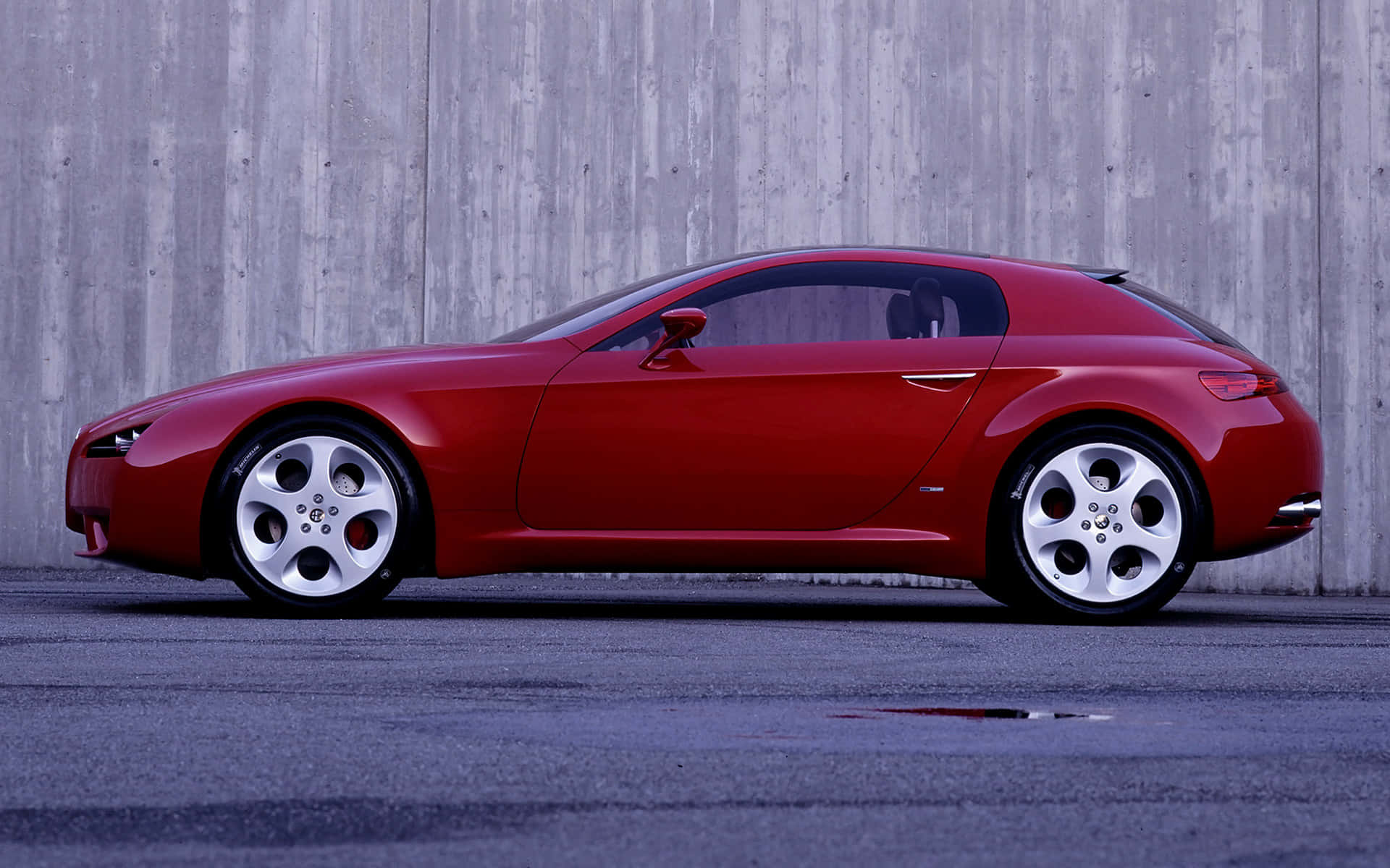 Alfa Romeo Brera 桌布 壁紙 — 一輛時尚的紅色跑車配備銀色輪圈，停放在風化的灰色混凝土牆旁，地面為鋪砌表面。