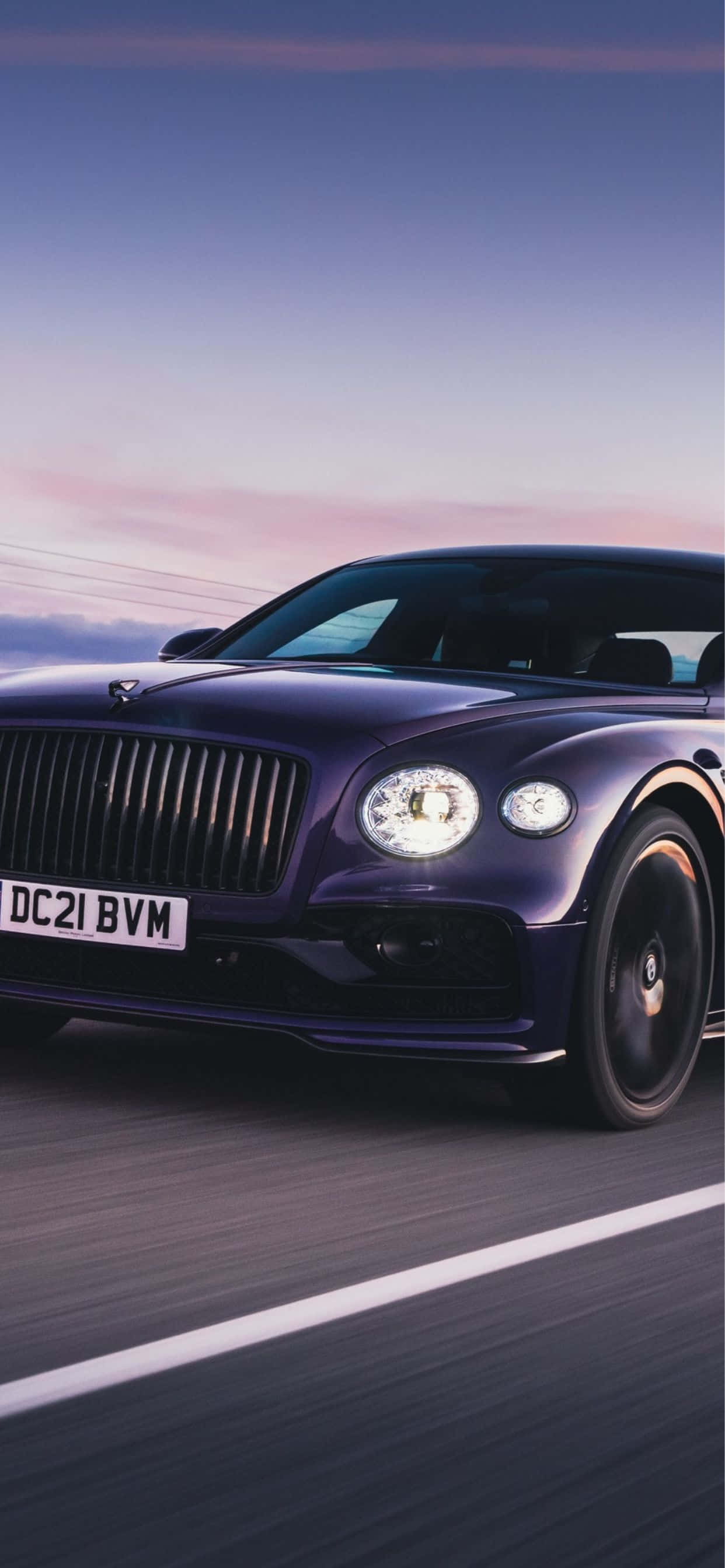 iPhone Xs Max Bentley 桌布 壁紙 — 一輛時尚的紫色賓利在黃昏的天空下，帶著柔和的粉紅色和藍色色調，沿著道路飛馳。