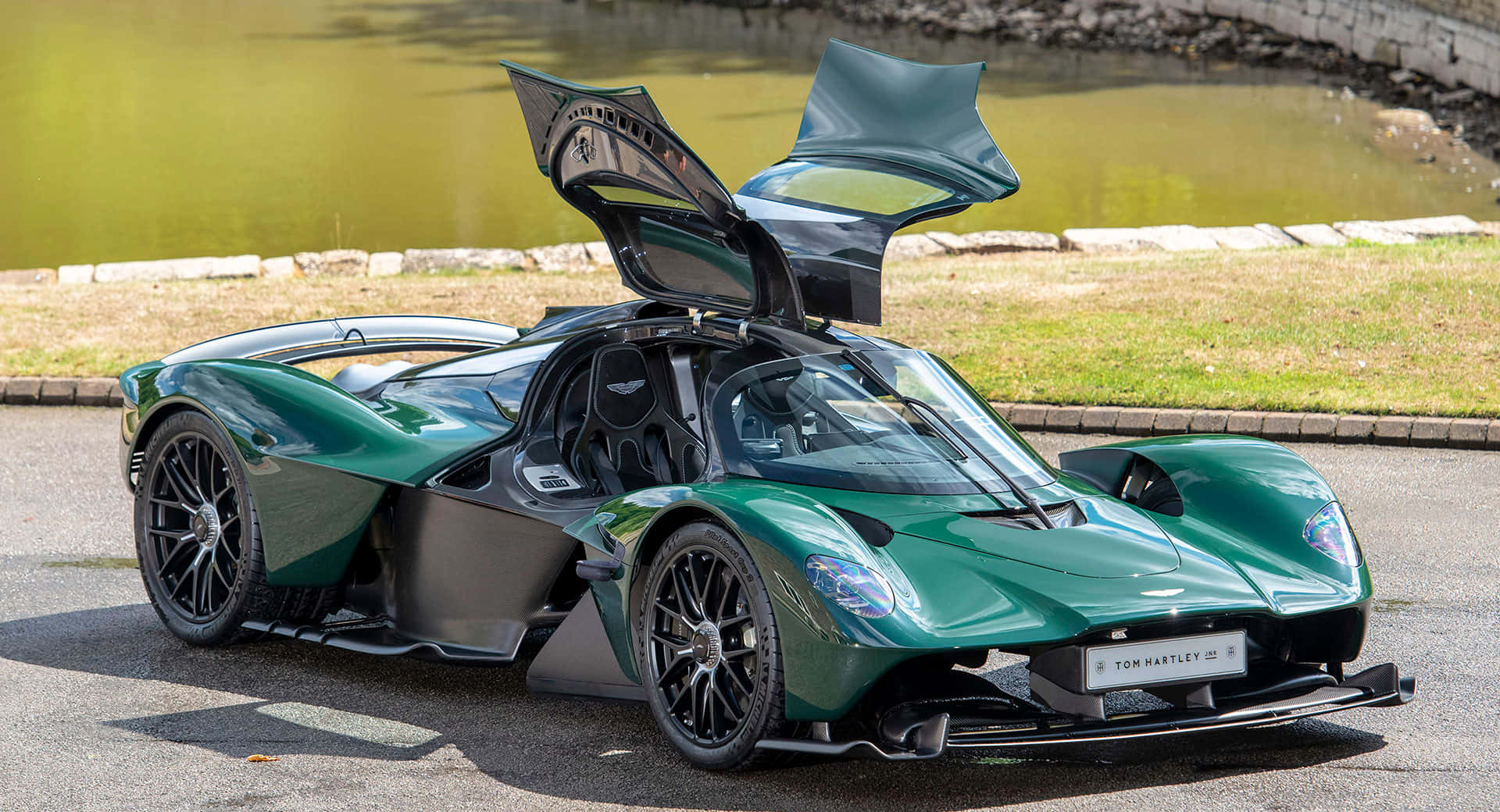 奧斯頓馬丁 桌布 壁紙 — 一輛時尚的綠色 Aston Martin Valkyrie 超跑，鷗翼車門打開，停泊在陽光明媚的池塘旁邊的瀝青路上。