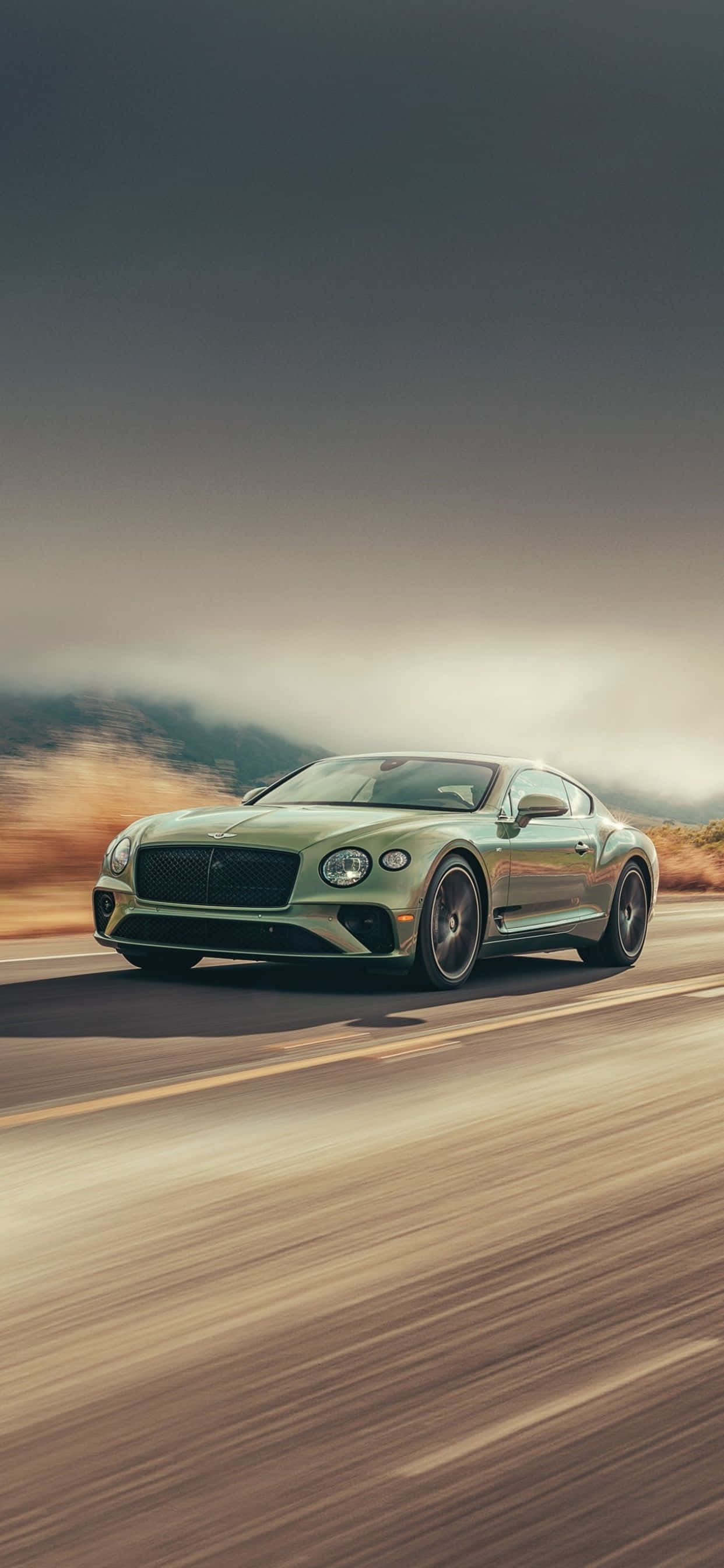 iPhone Xs Max Bentley 桌布 壁紙 — 一輛時尚的綠色 Bentley Continental GT 在沙漠公路上疾馳，背景是模糊的動態和朦朧的山巒。