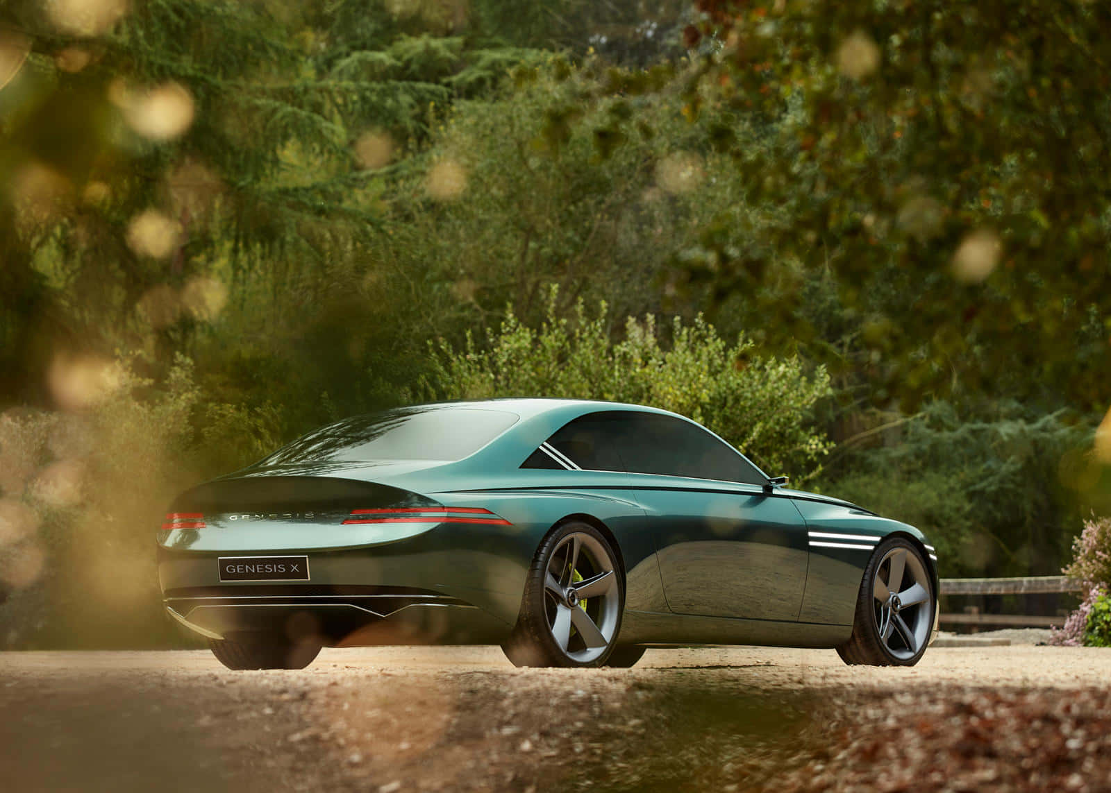 Genesis X Concept 桌布 壁紙 — 一輛時尚的綠色 Genesis X 概念車停在碎石路上，前景有模糊的森林樹葉和金色散景。