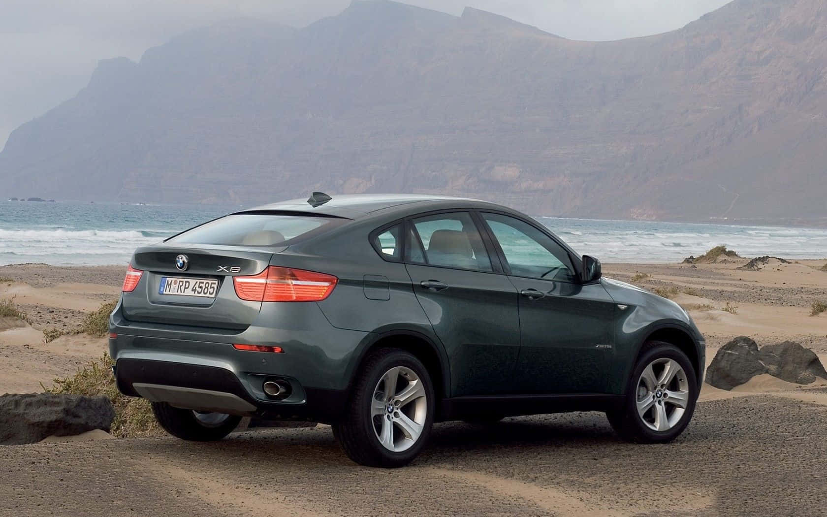 Bmw X6 桌布 壁紙 — 一輛時尚的綠色BMW X6 SUV停放在崎嶇的海岸沙灘上，背景中海浪拍打著懸崖。