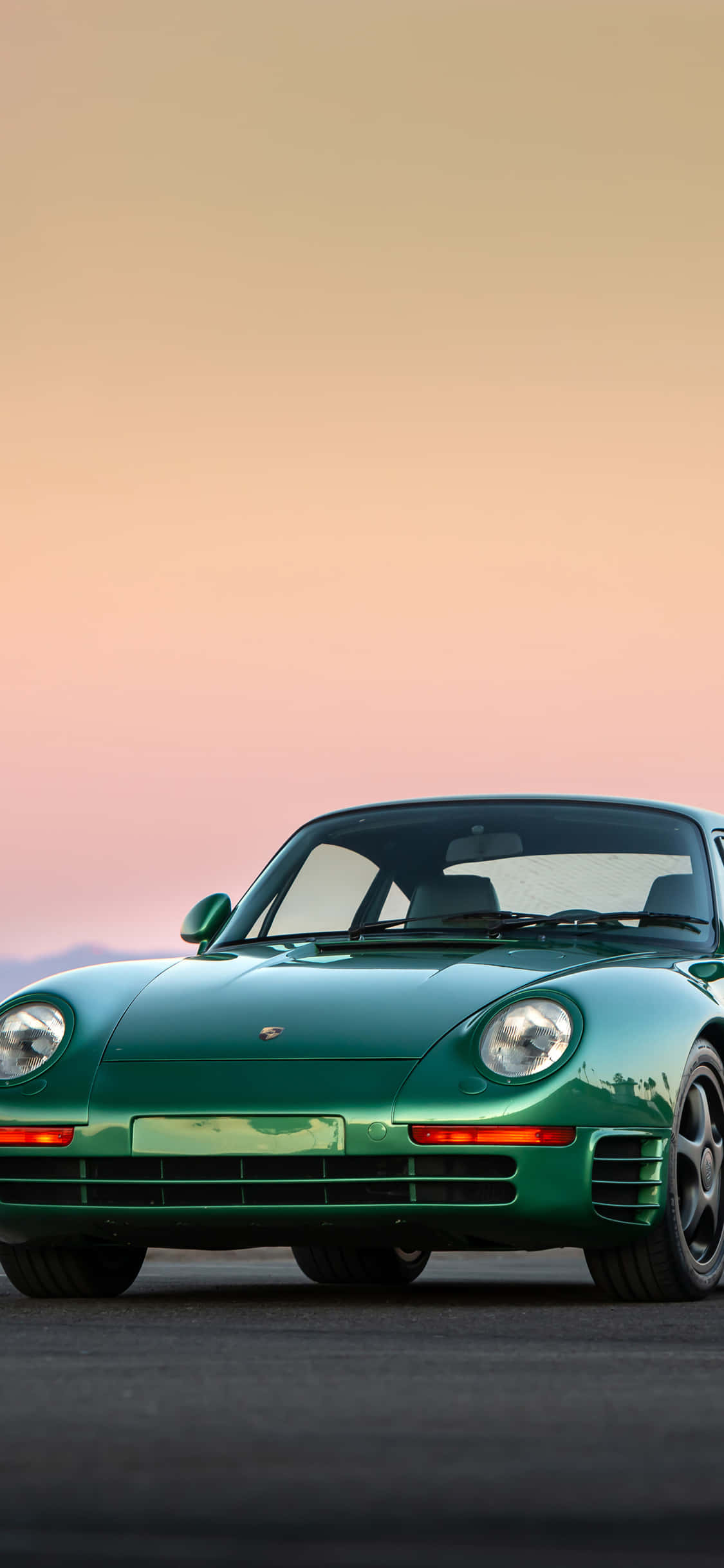 Porsche iPhone 桌布 壁紙 — 一輛時尚的綠色保時捷959跑車停在瀝青路面上，背景是柔和的粉紅與橙色日落天空。
