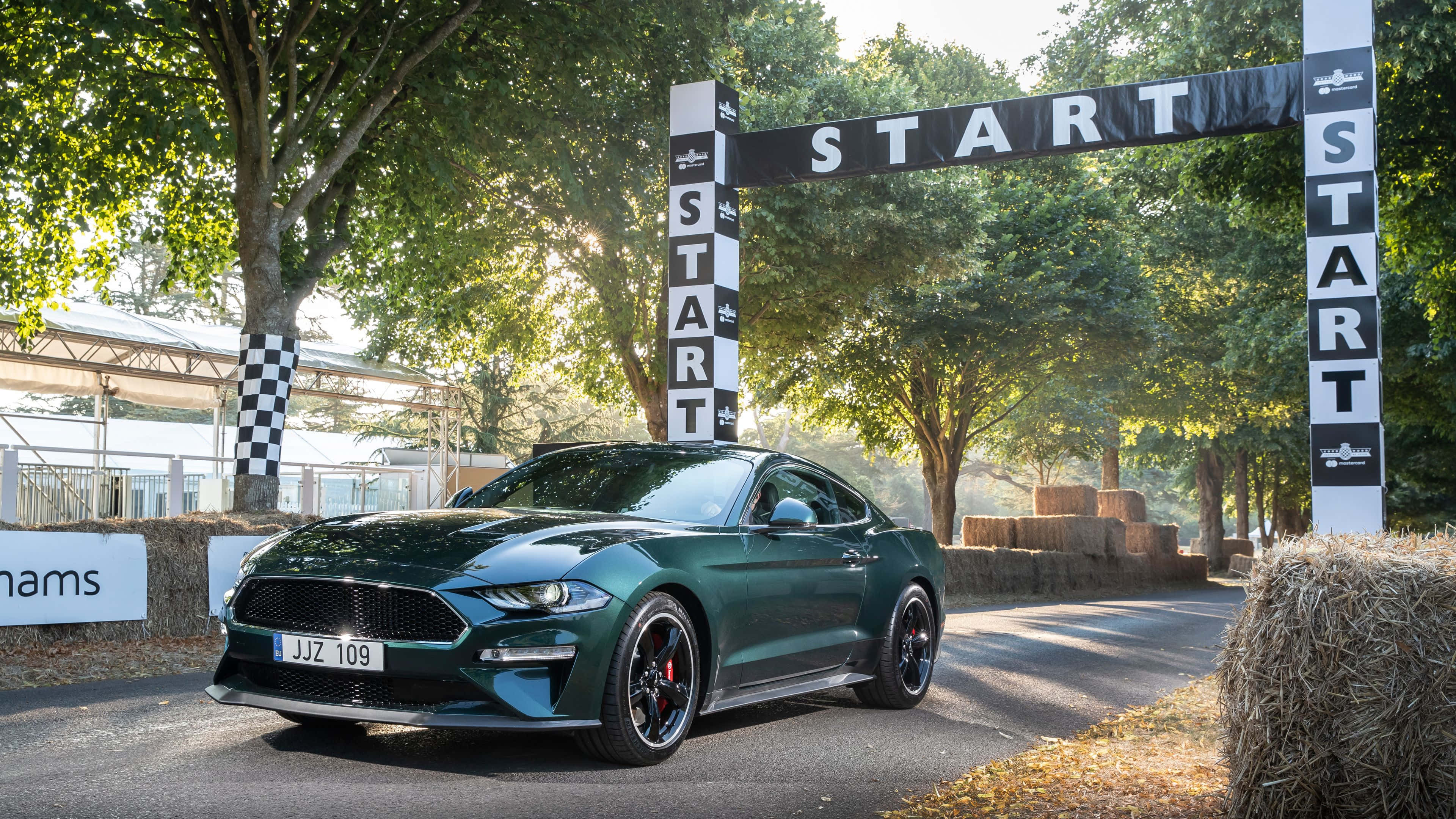 Ford Mustang Bullitt 桌布 壁紙 — 一輛時尚的綠色福特野馬停在賽車起跑線上，下方是黑白拱門，周圍環繞著樹木和乾草捆。