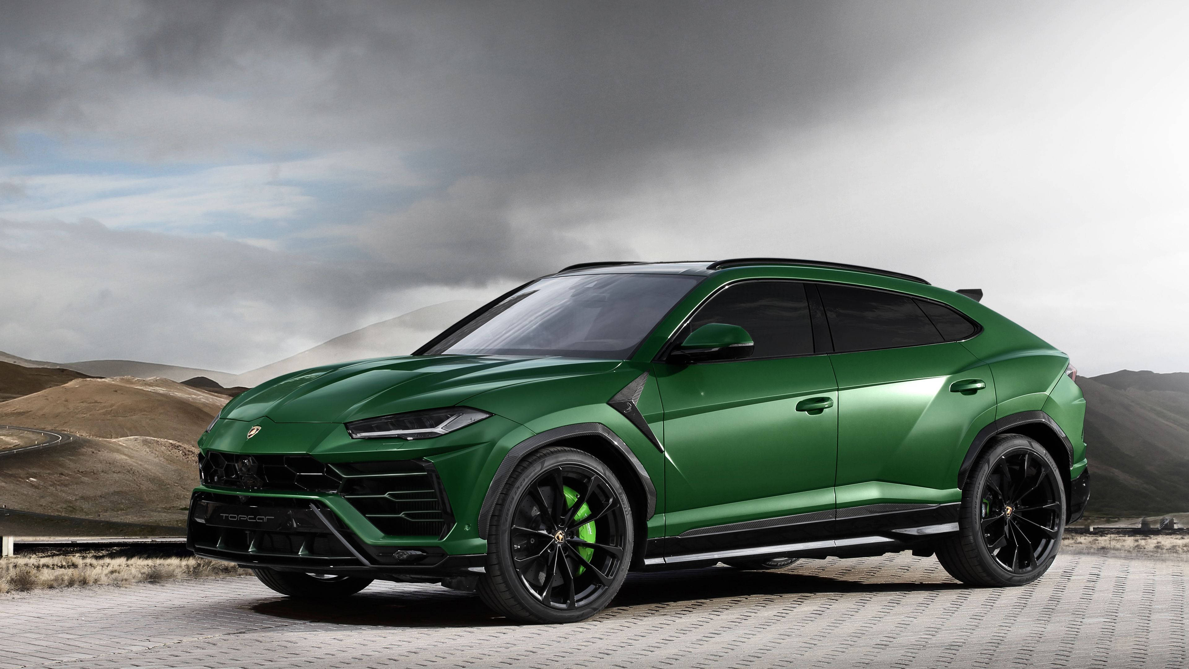 lambo 桌布 壁紙 — 一輛時尚的綠色藍寶堅尼 Urus 停在鋪砌的道路上，背景是戲劇性的多雲天空和起伏的山丘。