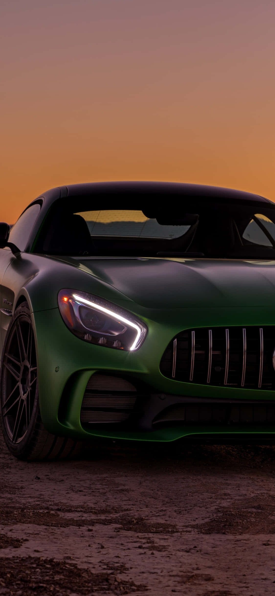 iPhone X Amg Gt-r 桌布 壁紙 — 一輛時尚的綠色跑車在溫暖的橙色日落天空下閃耀，車頭燈發出光芒。