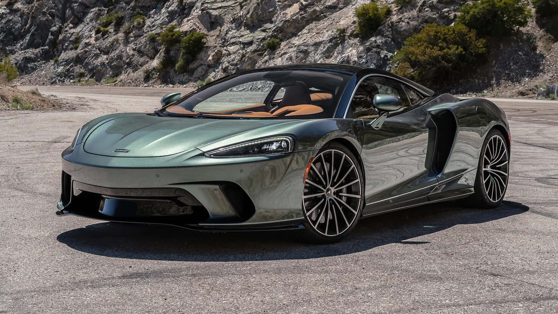 麥拉倫 桌布 壁紙 — 一輛時尚的綠色金屬McLaren跑車停泊在陽光照耀的瀝青路上，背景有岩石峭壁和稀疏的植被。