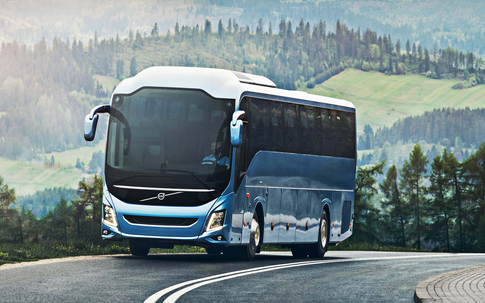 Bus 桌布 壁紙 — 一輛時尚的藍白色富豪長途巴士沿著蜿蜒的山路行駛，穿過茂密的綠色森林。