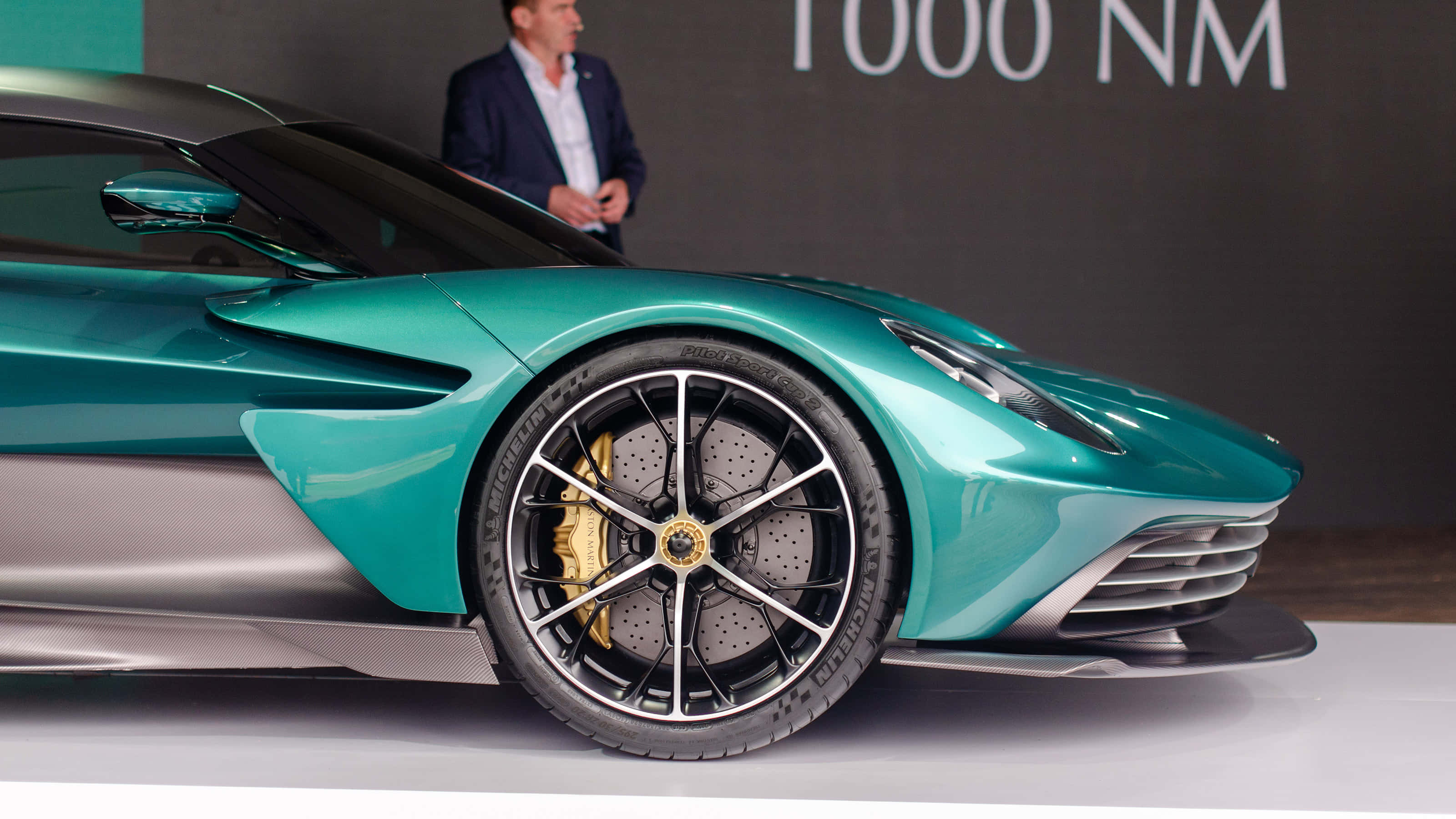 奧斯頓馬丁 桌布 壁紙 — 一輛時尚的藍綠色 Aston Martin 超級跑車配備金色制動卡鉗，在白色平台上於汽車活動中展出。