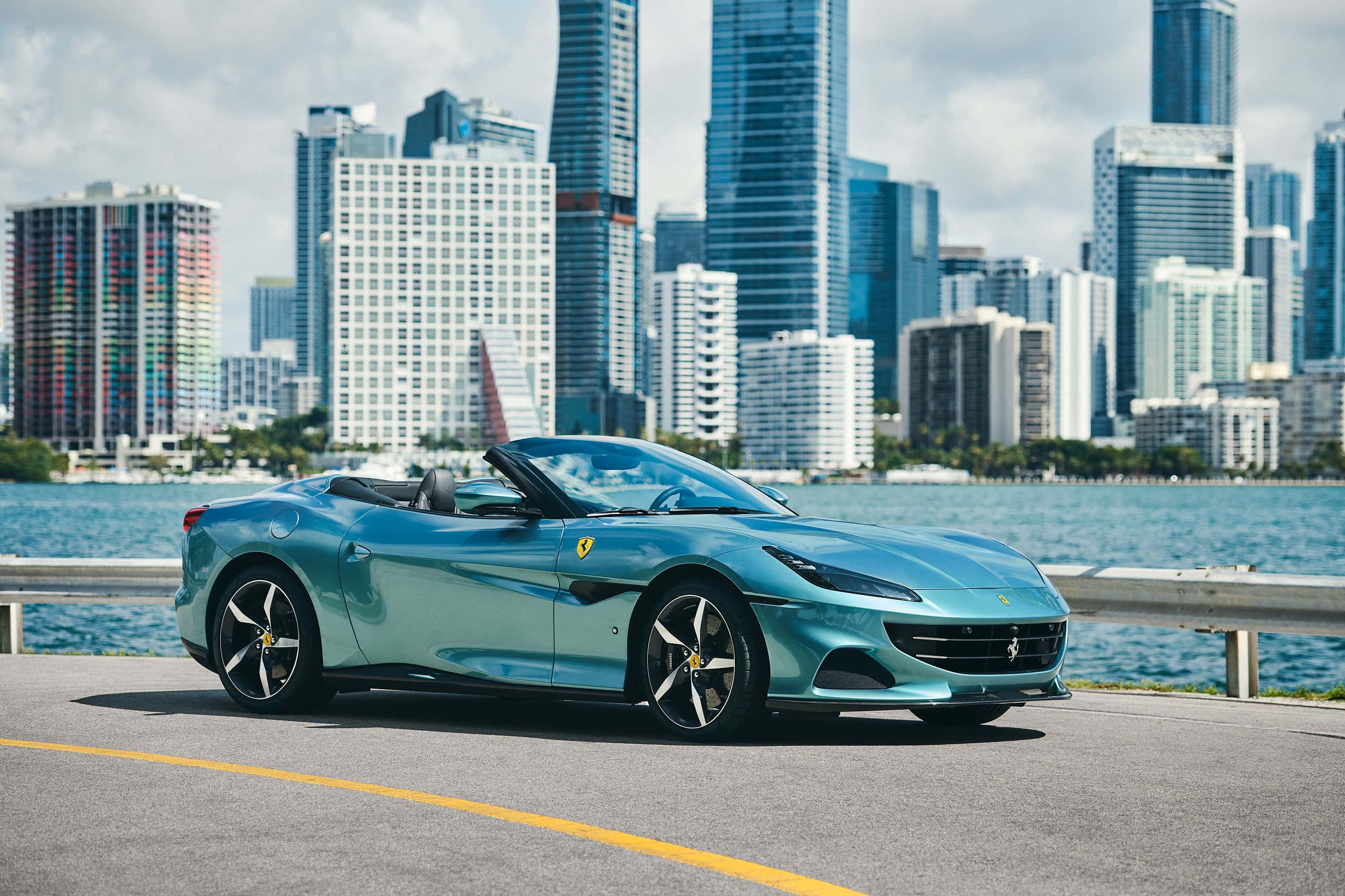 Ferrari Portofino 桌布 壁紙 — 一輛時尚的藍綠色法拉利敞篷車停在海濱道路上，背後是現代城市天際線和藍色海水。