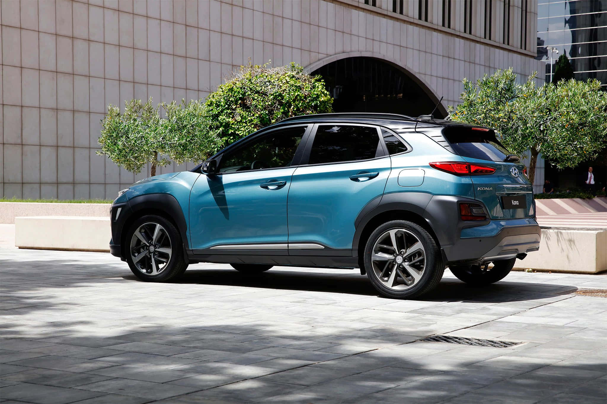 Hyundai 桌布 壁紙 — 一輛時尚的藍綠色現代Kona SUV停放在陽光普照的石頭廣場上，旁邊是現代建築和綠色灌木。