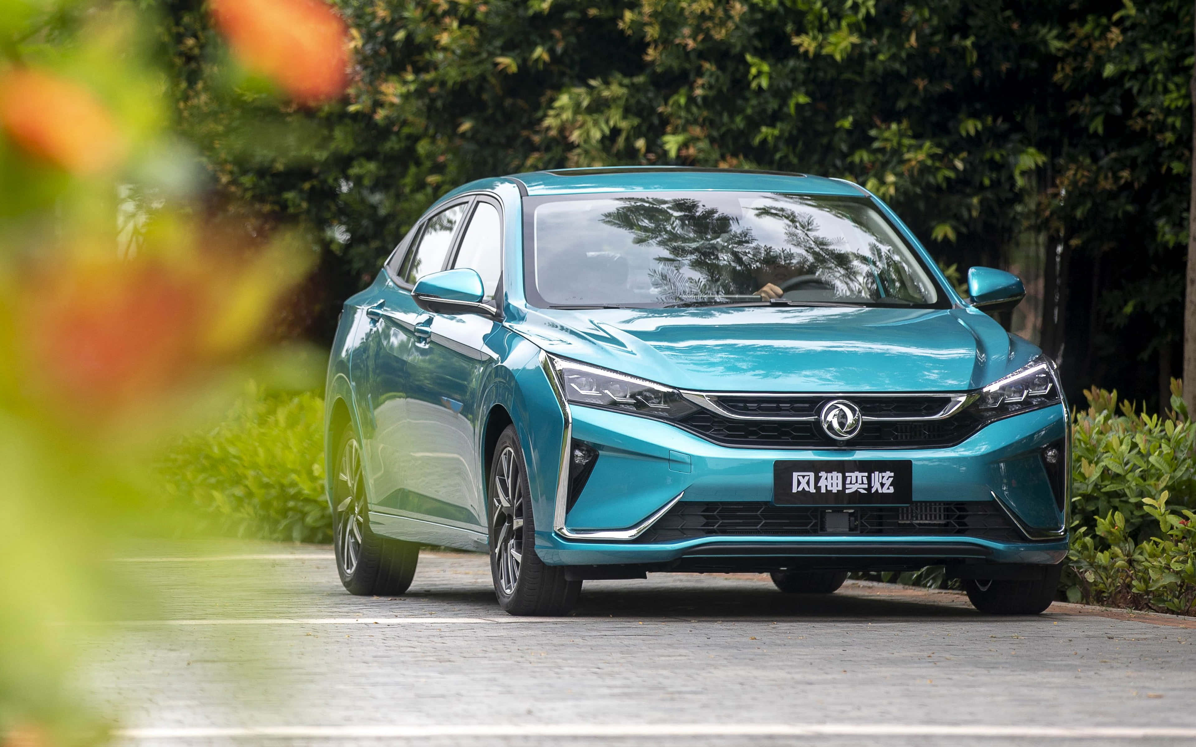 Dongfeng 桌布 壁紙 — 一輛時尚的藍綠色轎車停放在鋪砌的小路上，周圍環繞著茂密的綠色樹葉和模糊的橙色花朵。
