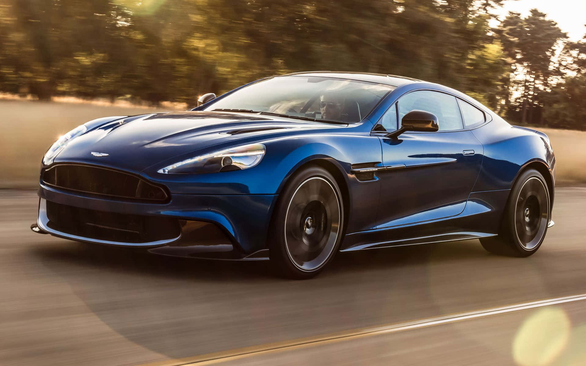 Aston Martin Vanquish 桌布 壁紙 — 一輛時尚的藍色 Aston Martin 跑車在陽光明媚的道路上行駛，背景是模糊的樹木。