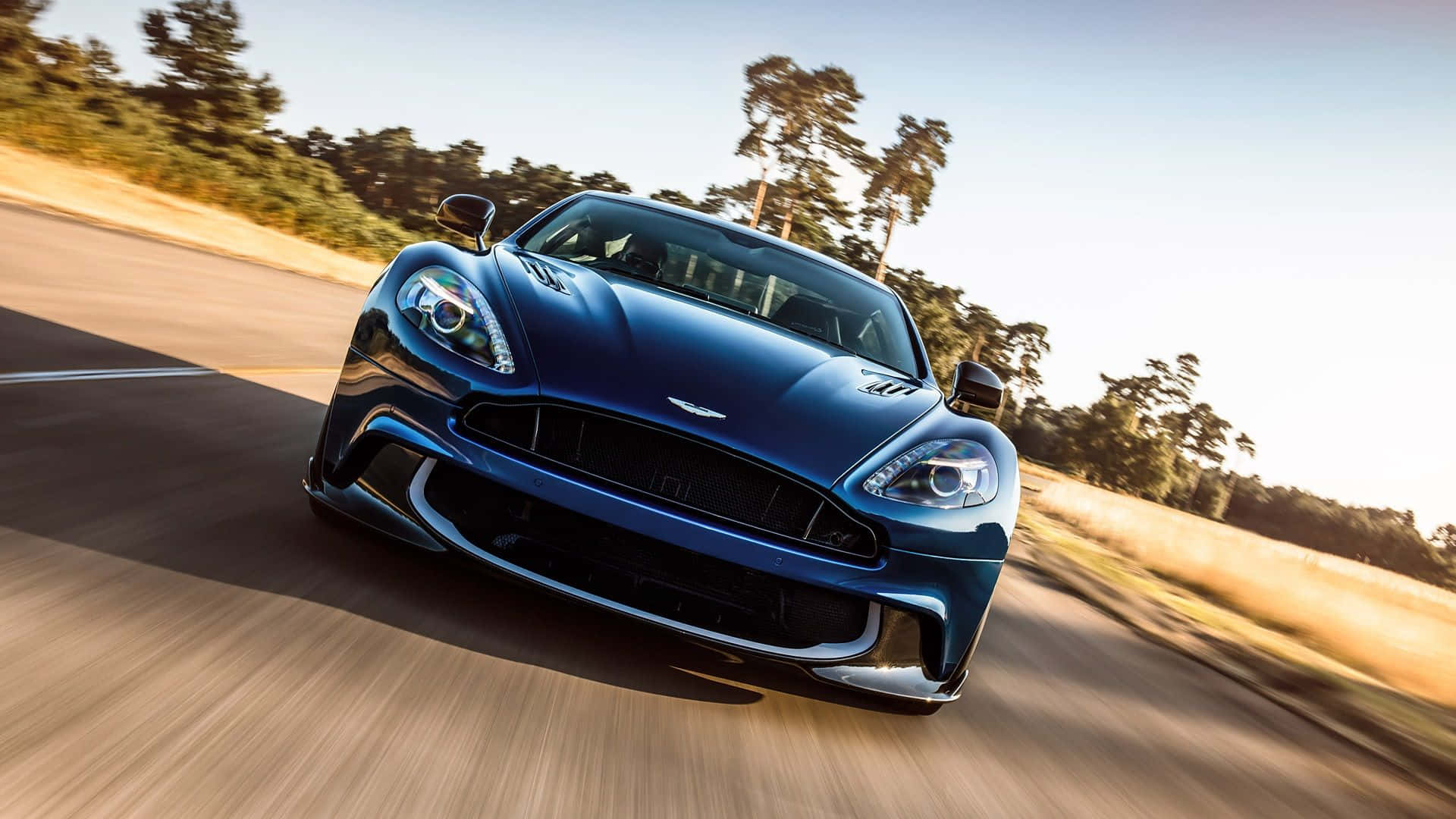 Aston Martin Vanquish 桌布 壁紙 — 一輛時尚的藍色 Aston Martin 跑車在陽光明媚的鄉村道路上疾馳，背景有模糊的動感和樹木。