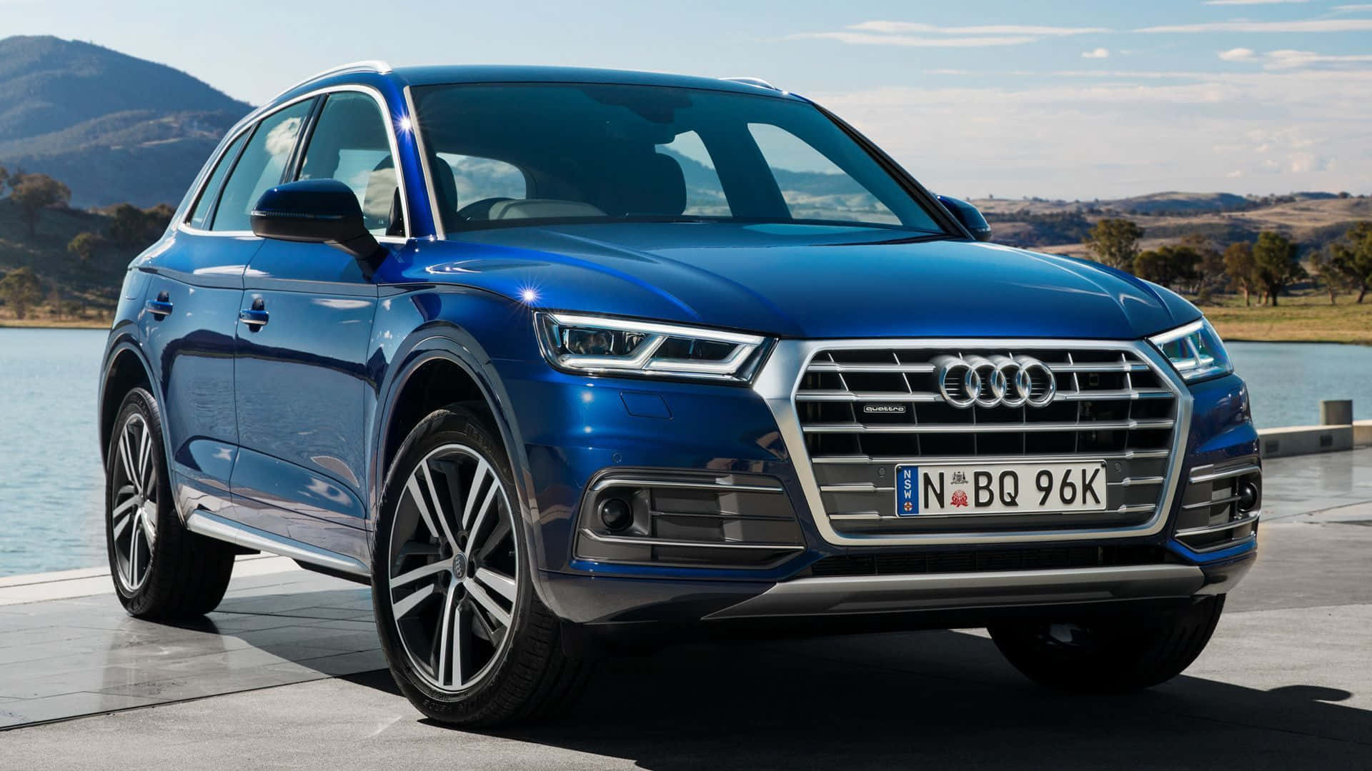 Audi Sq5 桌布 壁紙 — 一輛時尚的藍色 Audi Q5 SUV 停在平靜的湖邊，背景是起伏的丘陵和明亮的天空。