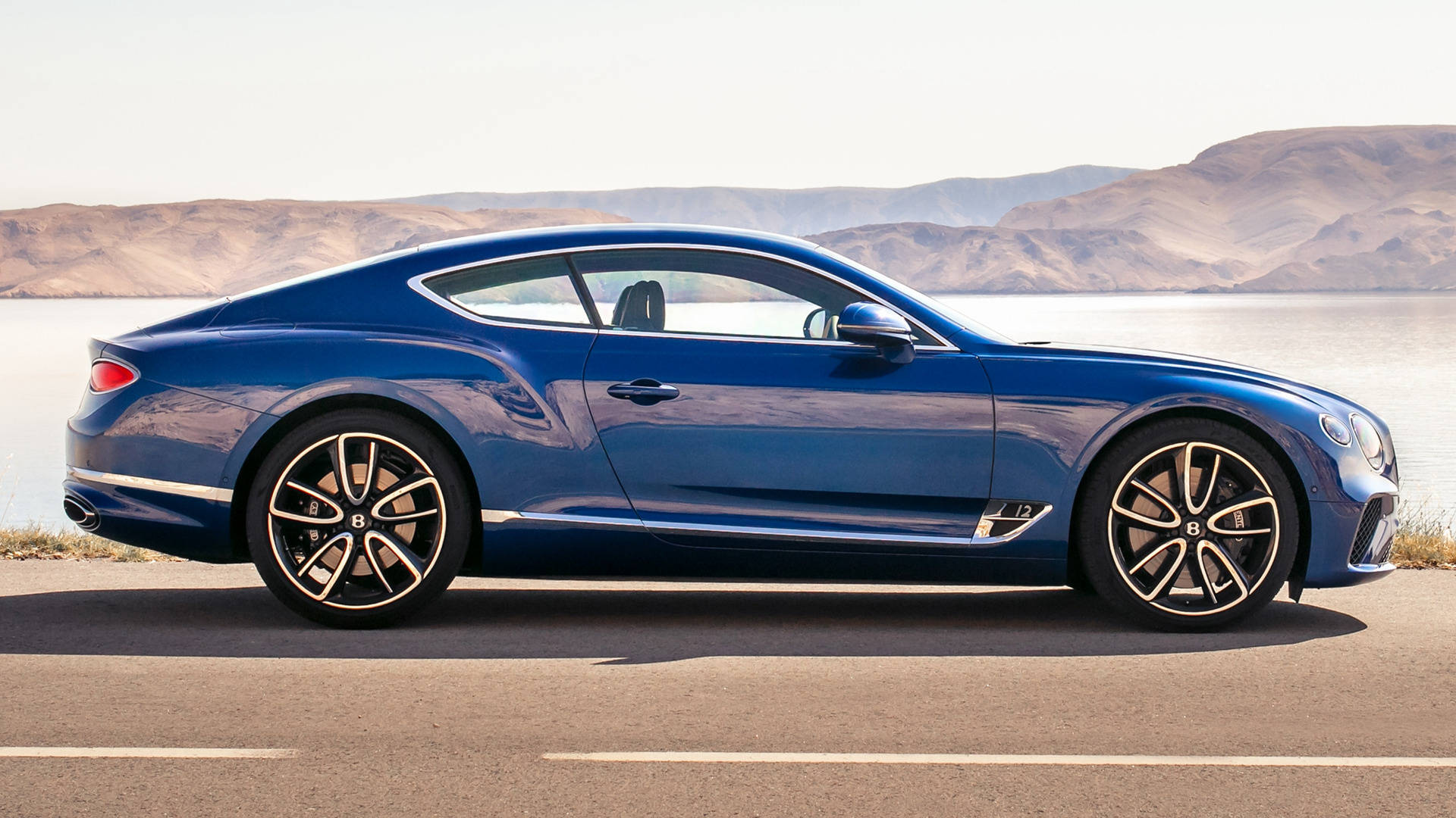 bentley HD 桌布 壁紙 — 一輛時尚的藍色 Bentley Continental GT 雙門跑車停泊在平靜的湖邊，背景是乾旱的山丘，在明亮的日光下。