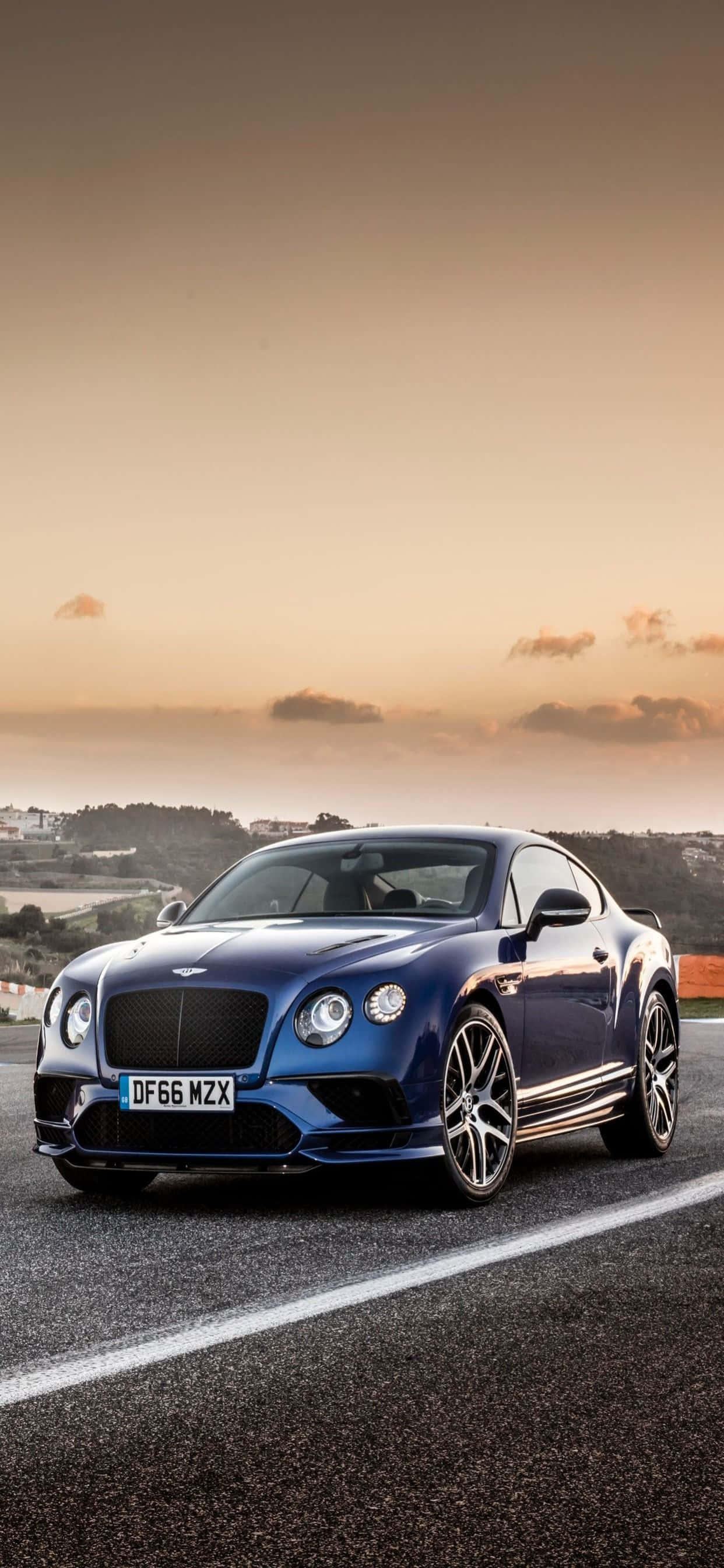 iPhone Xs Max Bentley 桌布 壁紙 — 一輛時尚的藍色 Bentley Continental GT 在日落時分沿著海岸公路奔馳，上方有溫暖的金色雲彩。