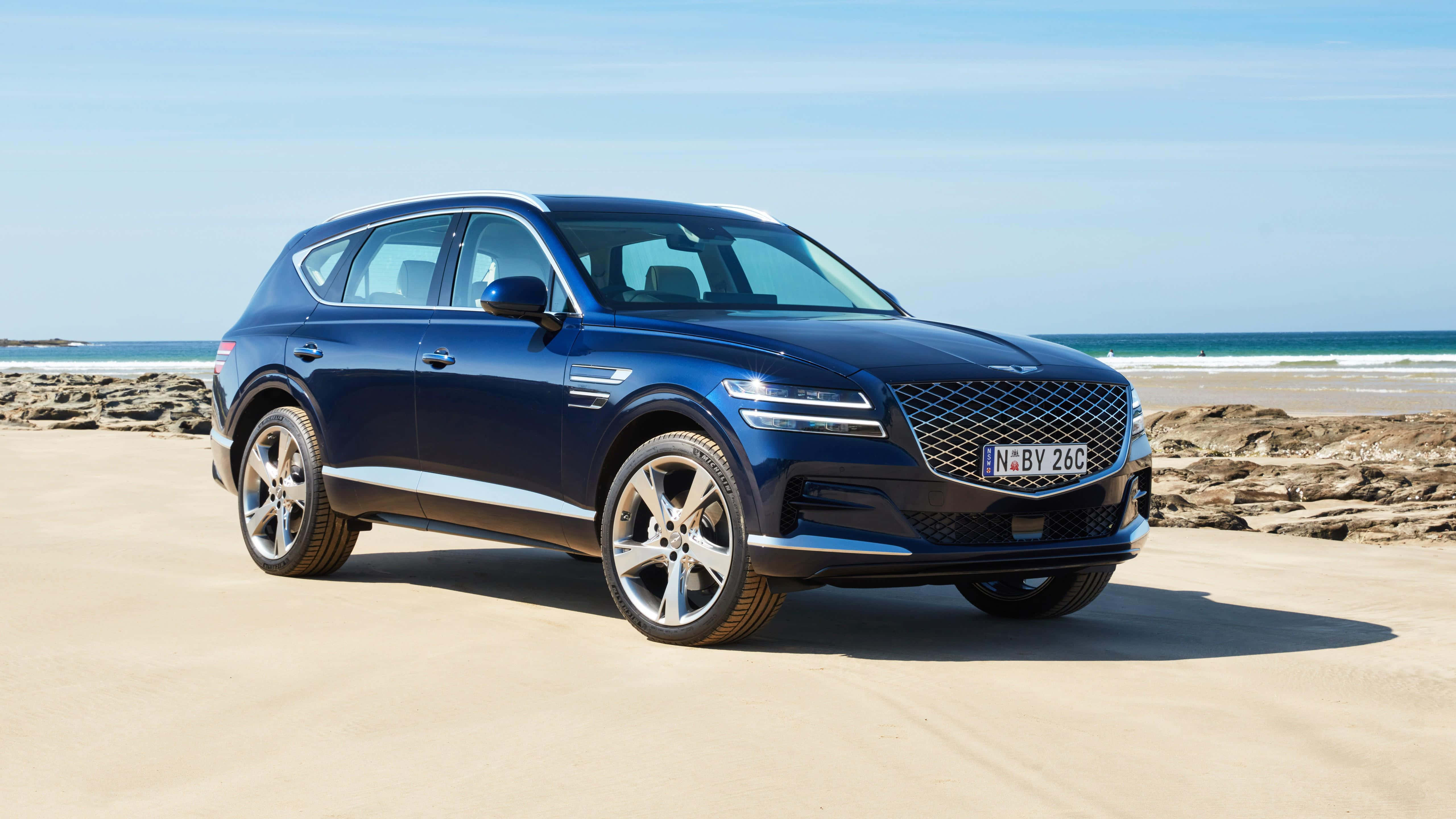 Genesis Essentia 桌布 壁紙 — 一輛時尚的藍色 Genesis GV80 SUV 停泊在陽光明媚的沙灘上，背景是海浪和岩石海岸。
