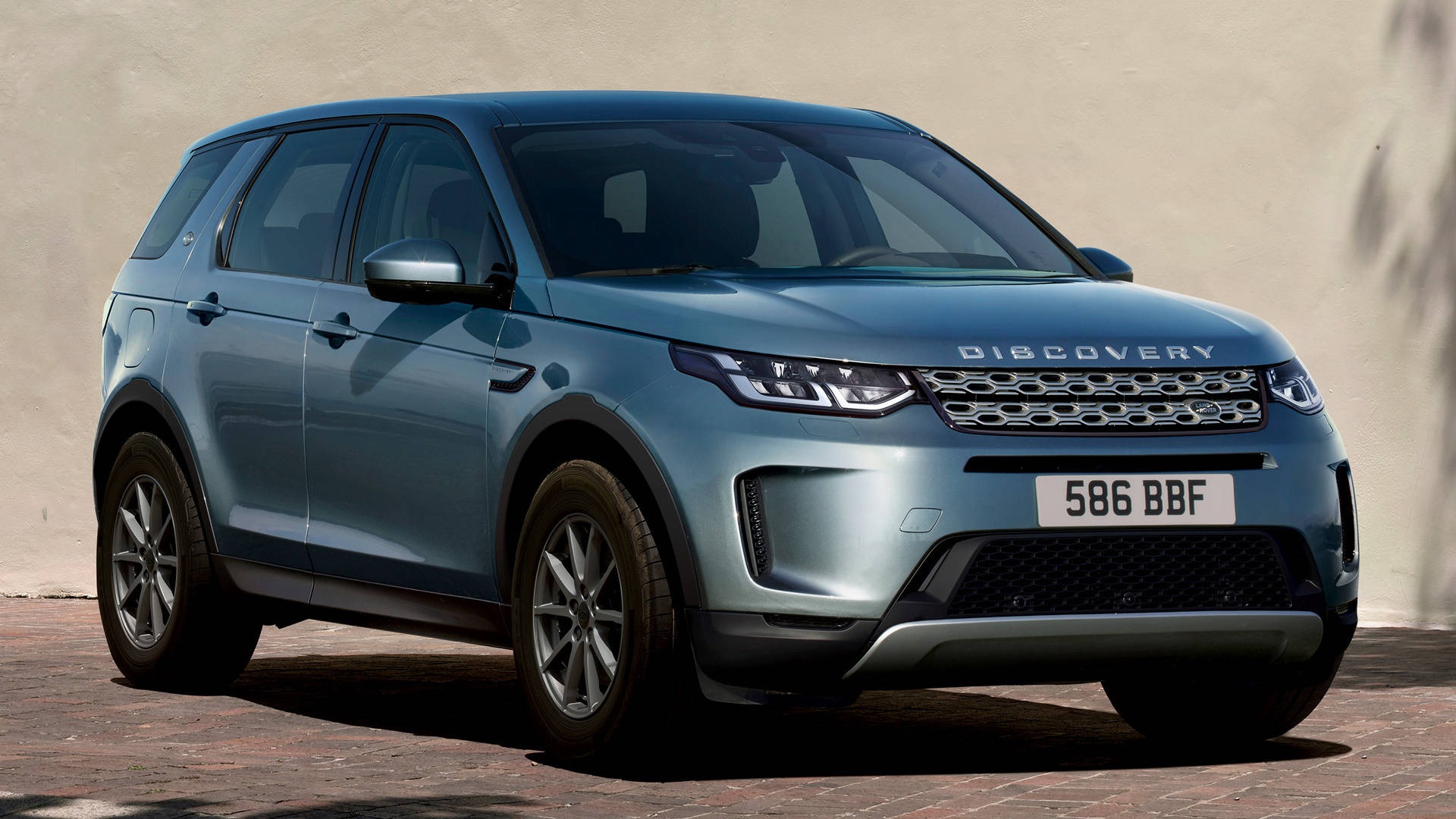 land rover iPhone 桌布 壁紙 — 一輛時尚的藍色 Land Rover Discovery SUV 停放在磚砌路面上，背景是米色牆壁，在明亮的日光下。
