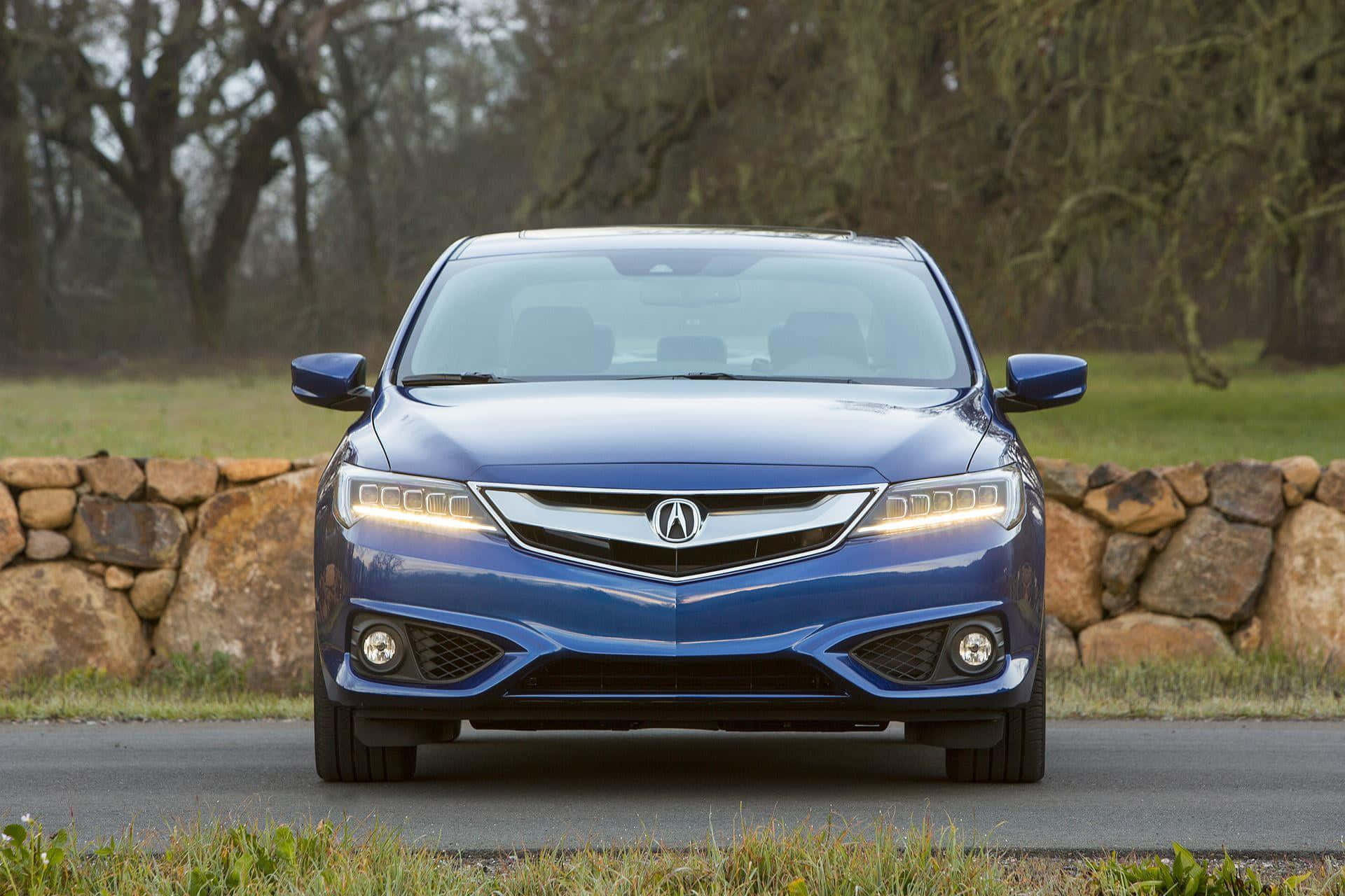 Acura Ilx 桌布 壁紙 — 一輛時尚的藍色Acura轎車在由石牆和光禿樹木環繞的柏油路上向前行駛。