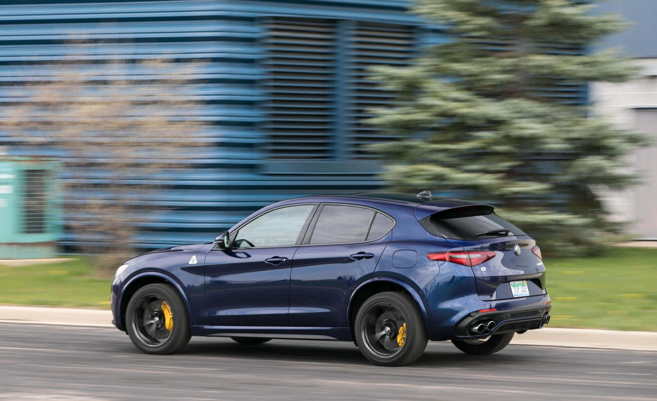 Alfa Romeo Stelvio 桌布 壁紙 — 一輛時尚的藍色Alfa Romeo Stelvio SUV在現代建築和綠植背景下高速駛過，帶有動態模糊效果。