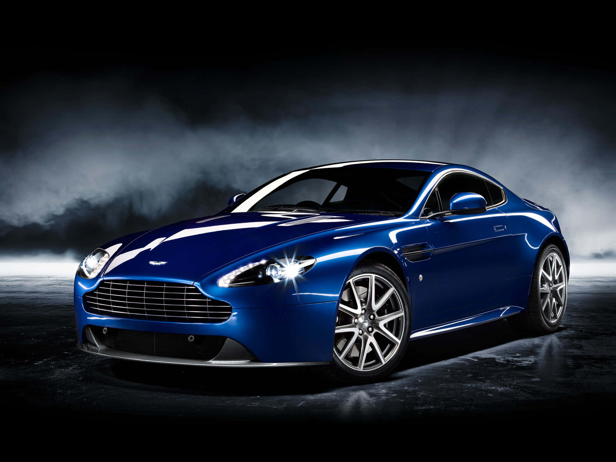 奧斯頓馬丁 桌布 壁紙 — 一輛時尚的藍色Aston Martin跑車在戲劇性的工作室燈光下，在煙霧瀰漫的黑暗背景中閃閃發光。