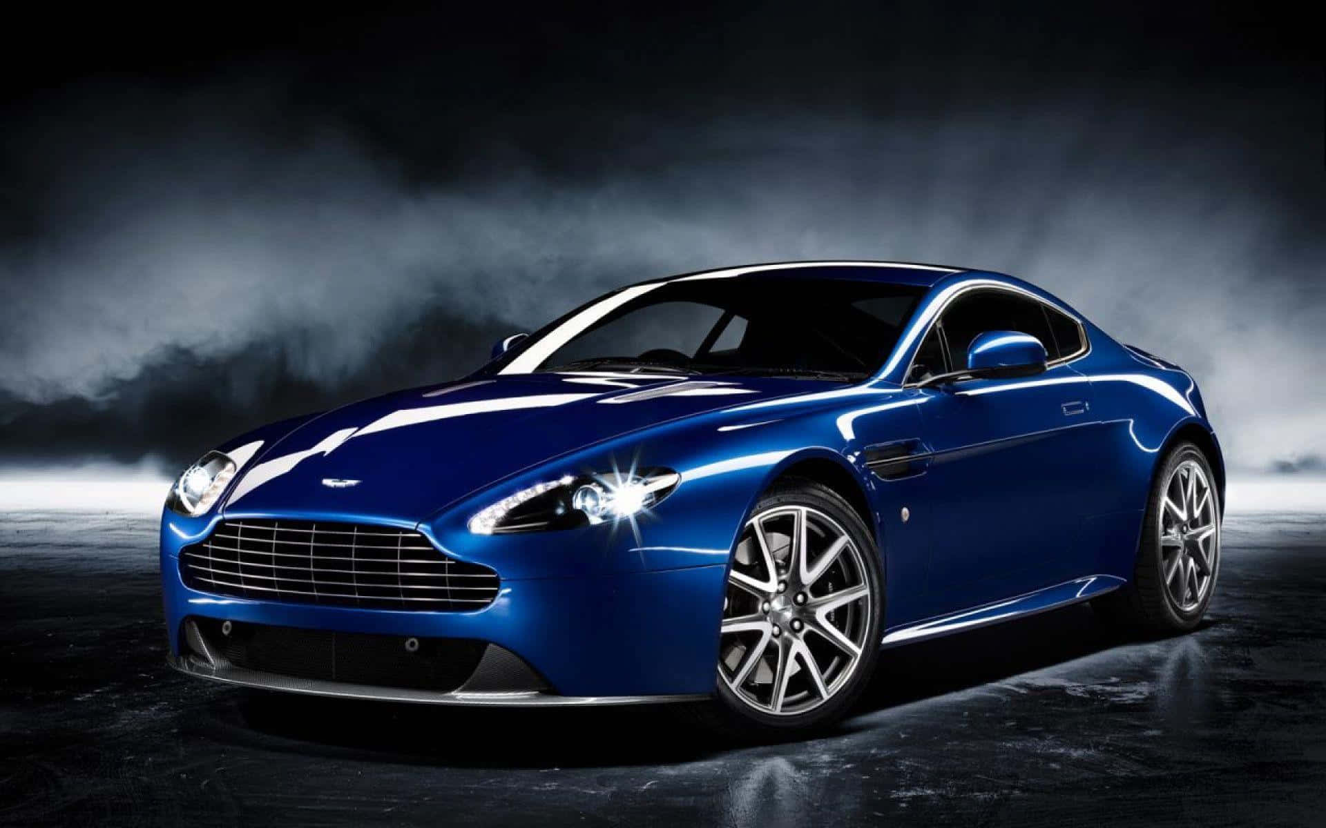Aston Martin Db9 桌布 壁紙 — 一輛時尚的藍色Aston Martin跑車在戲劇性的攝影棚燈光下，在煙霧瀰漫的黑暗背景中閃閃發光。