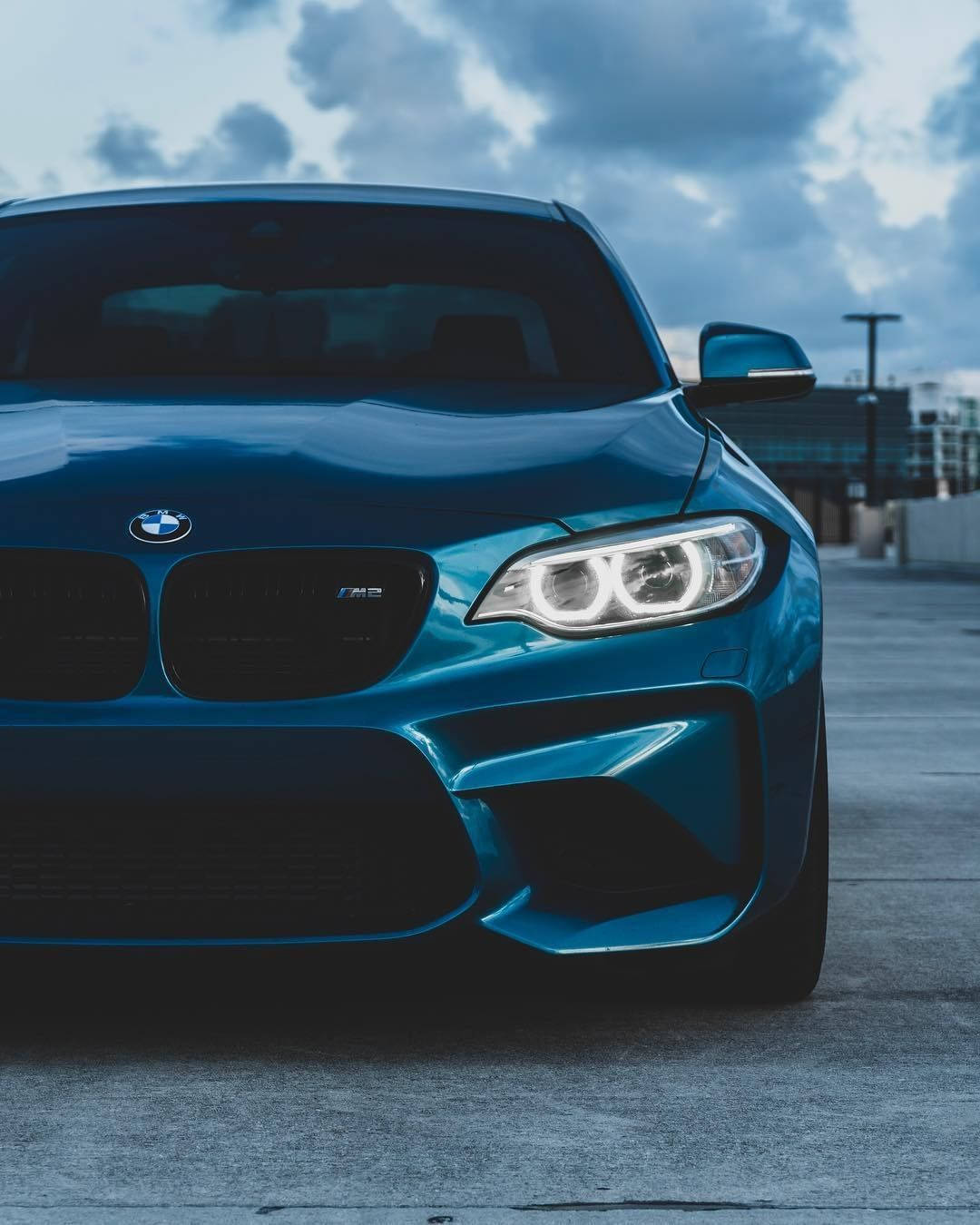 BMW 動圖 桌布 壁紙 — 一輛時尚的藍色BMW M2跑車，帶有發光的車頭燈，停放在多雲天空下的混凝土停車場。