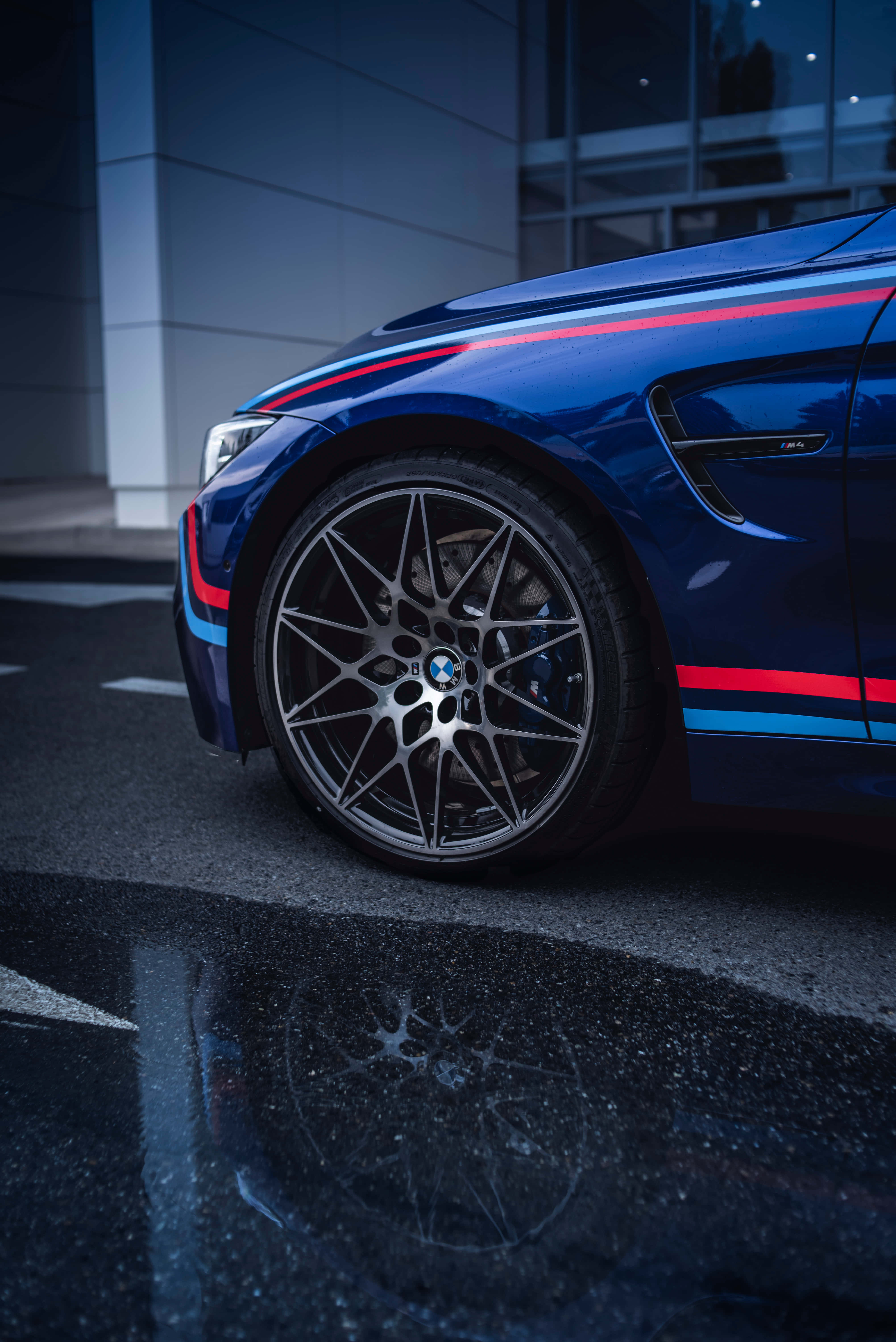 Best M Series 桌布 壁紙 — 一輛時尚的藍色BMW M4，帶有紅色和藍色賽車條紋，在暮色城市燈光下反射在濕漉漉的路面上。
